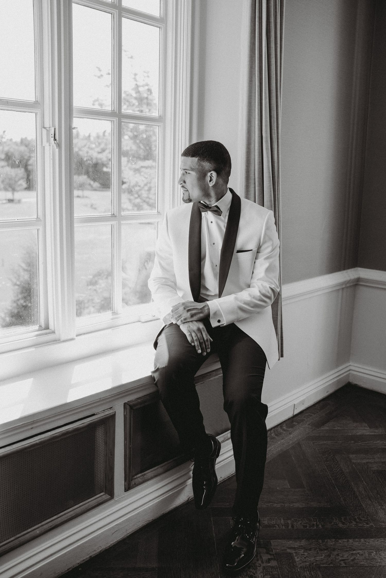 Groom in white tuxedo sitting on window sill in black and white photo.