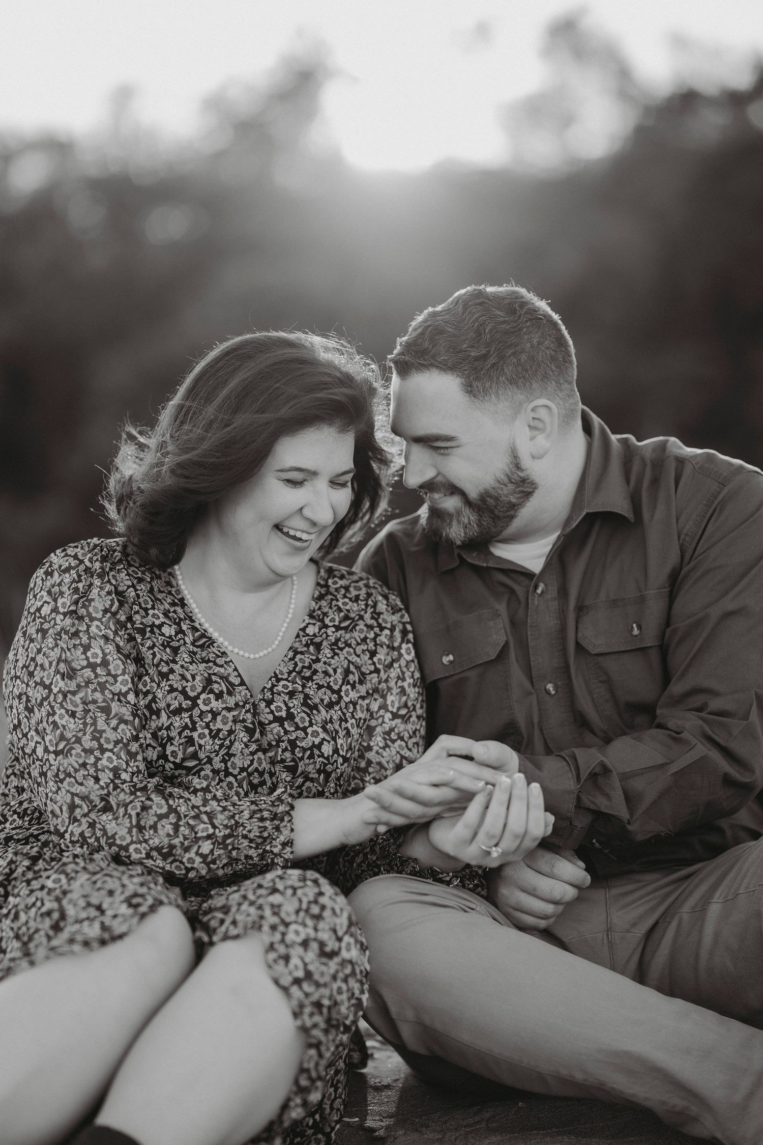Black and white photo of couple sitting together laughing and holding hands at sunset.