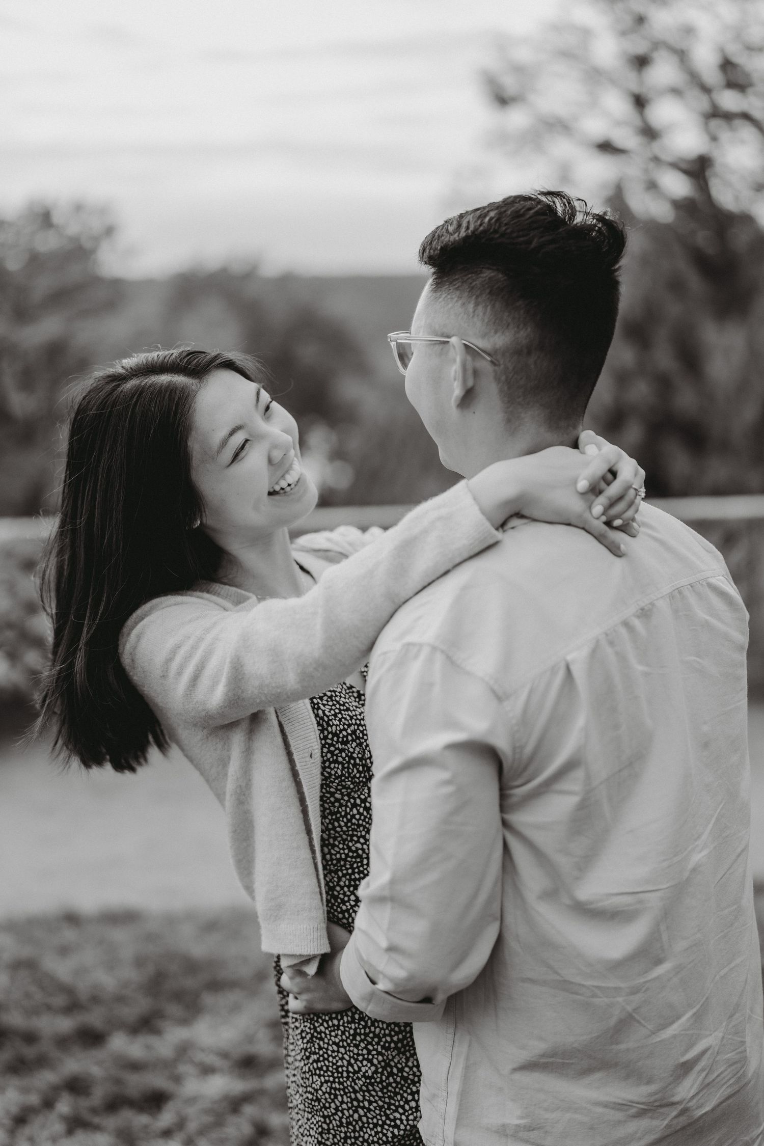 Black and white image of smiling woman embracing her partner outdoors during a romantic engagement session.