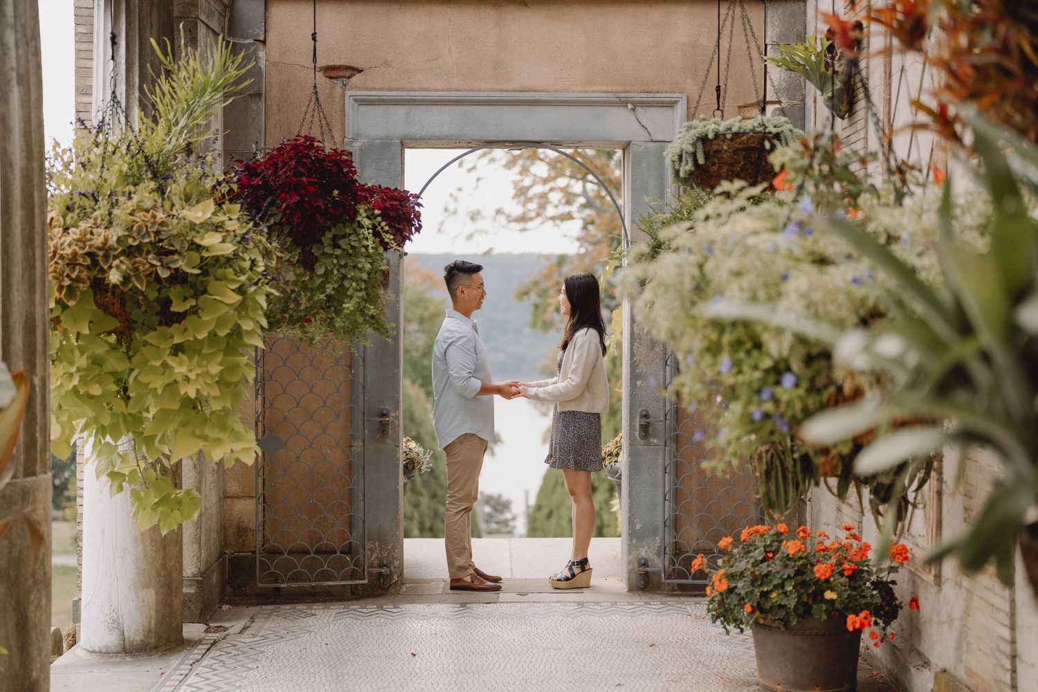 Couple holding hands in a stone garden archway surrounded by lush hanging plants and orange flowers for a romantic engagement photo session.
