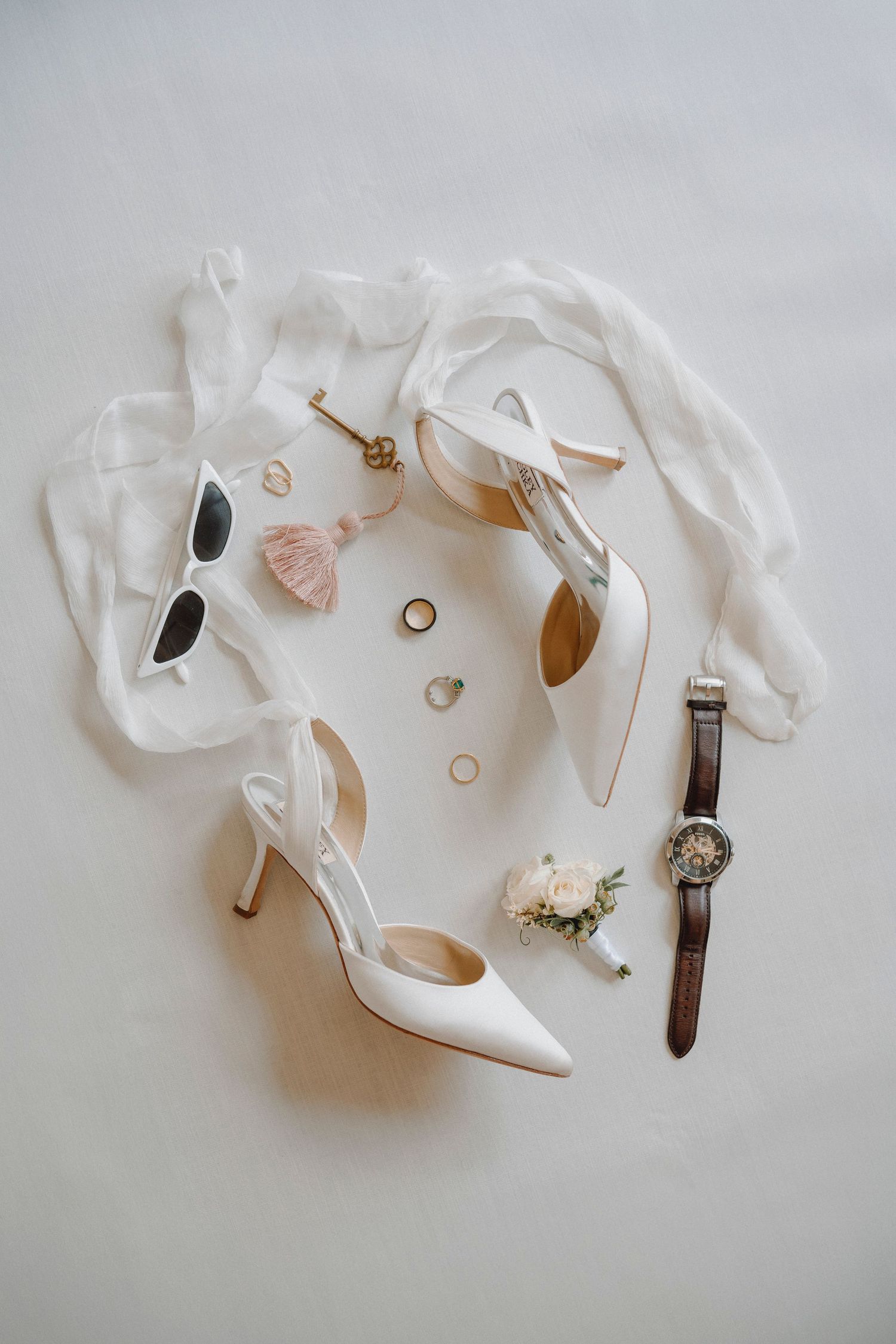 Flat lay of bridal accessories including white heels, rings, watch, sunglasses, and boutonniere.