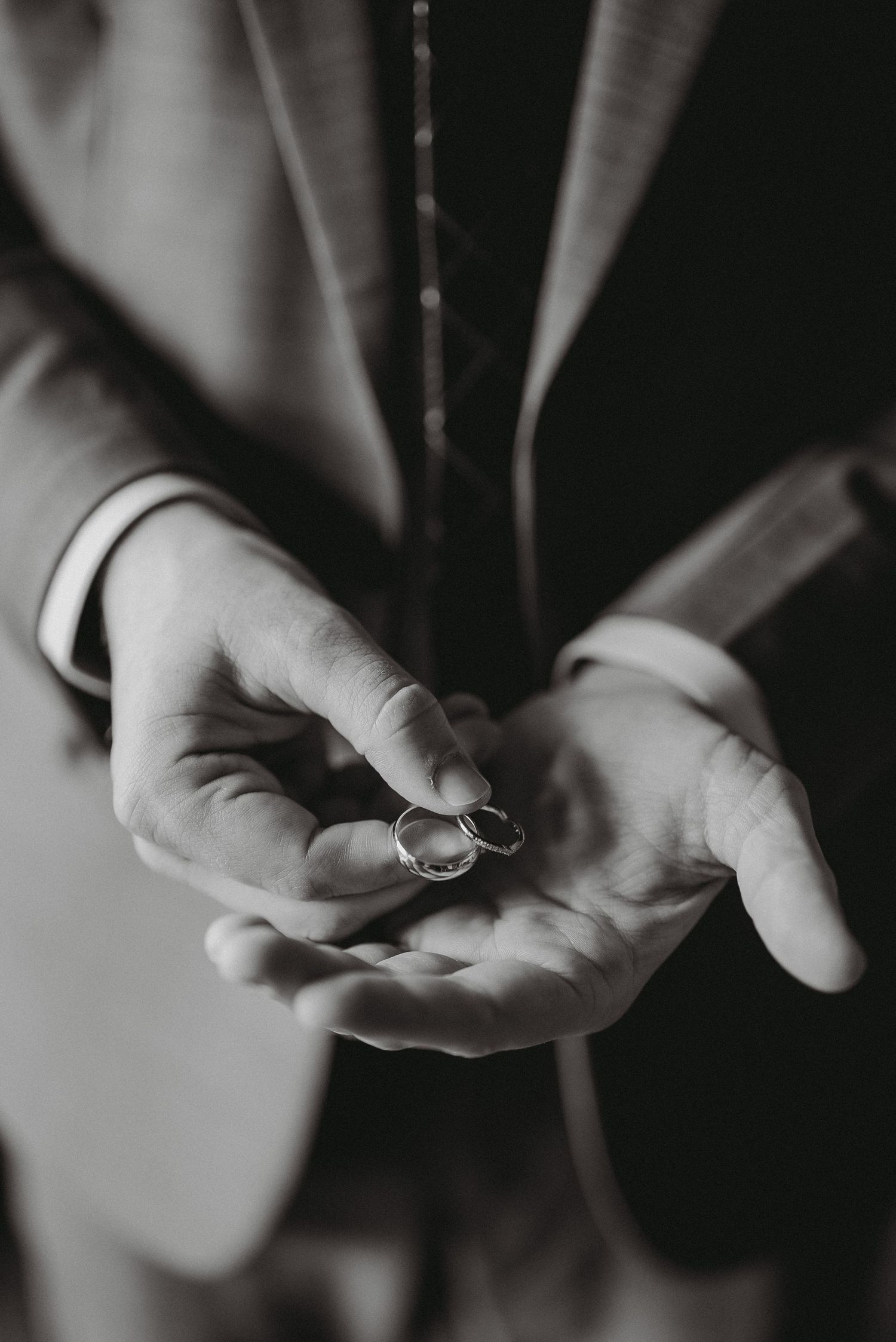 Close-up black and white photo of groom holding two wedding rings in open palms.