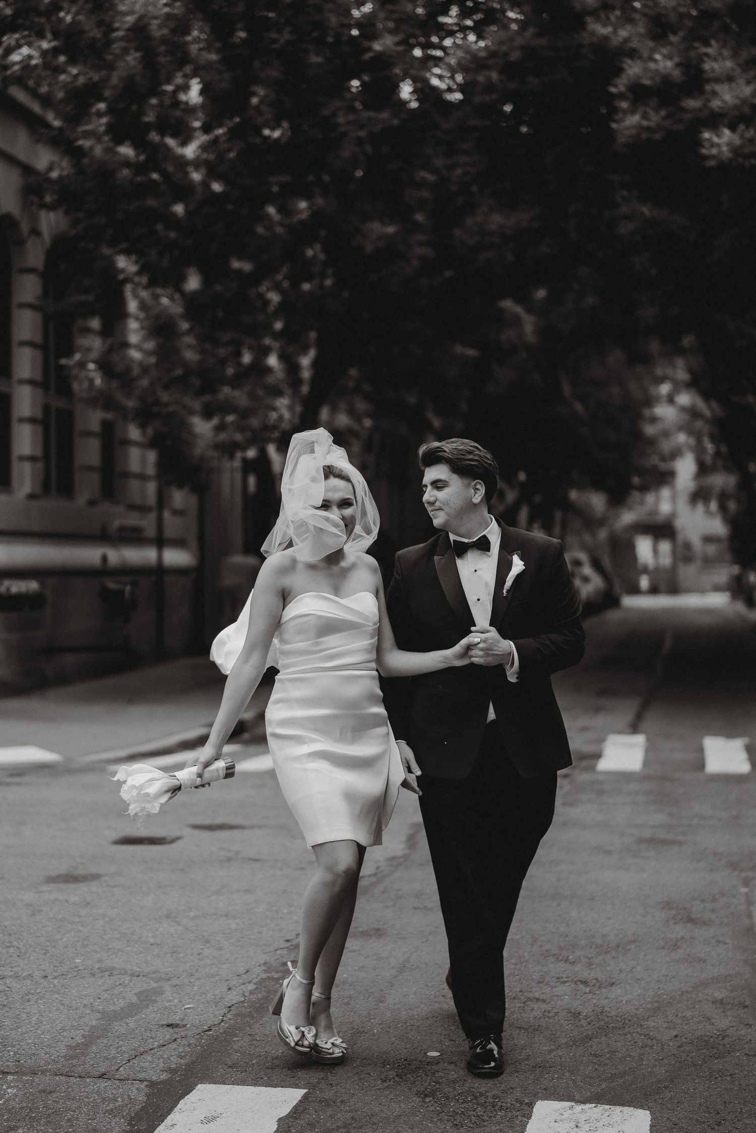 Black and white photo of bride and groom walking arm in arm across city crosswalk.