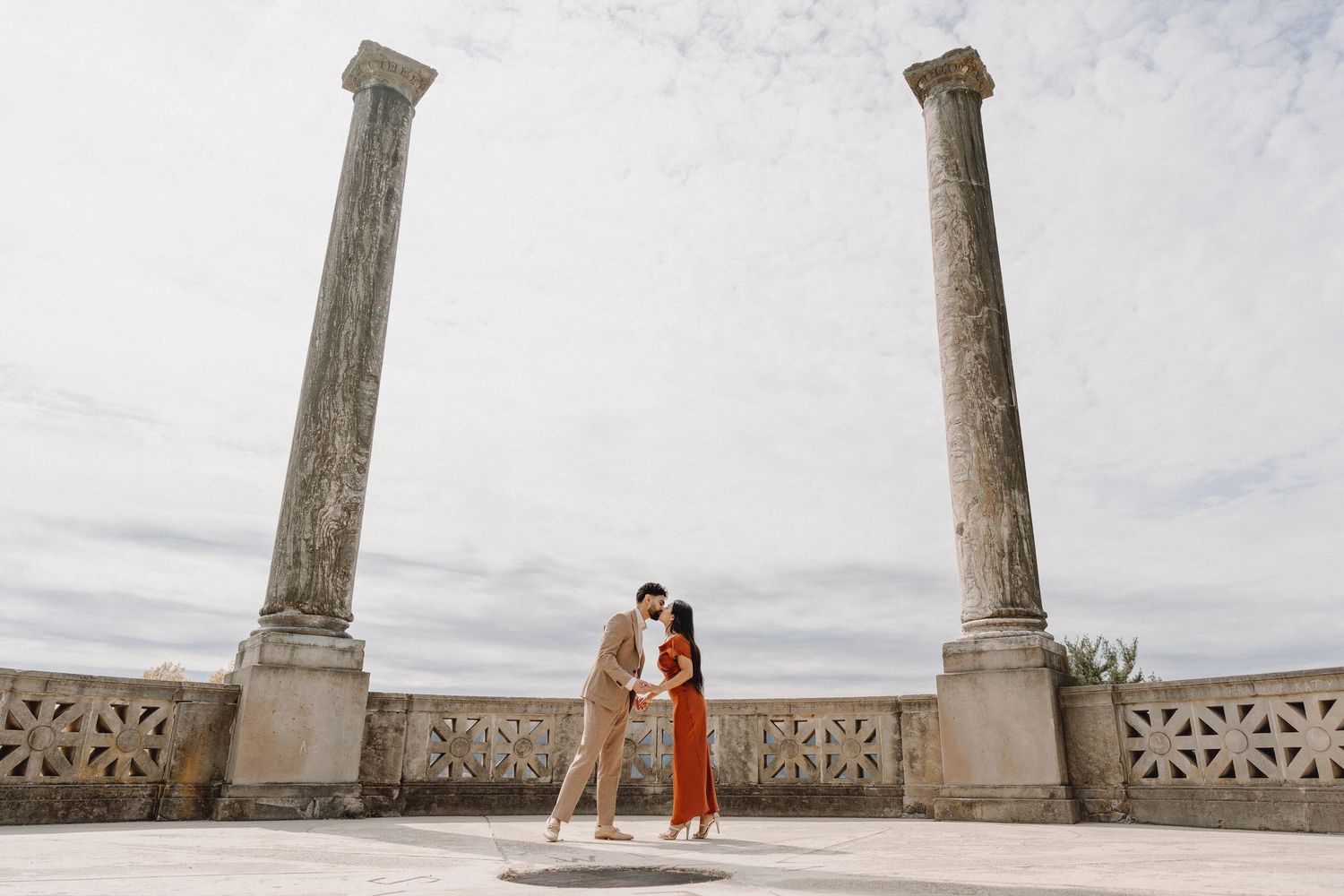 Couple sharing a kiss between two tall stone columns on an open terrace under cloudy skies.