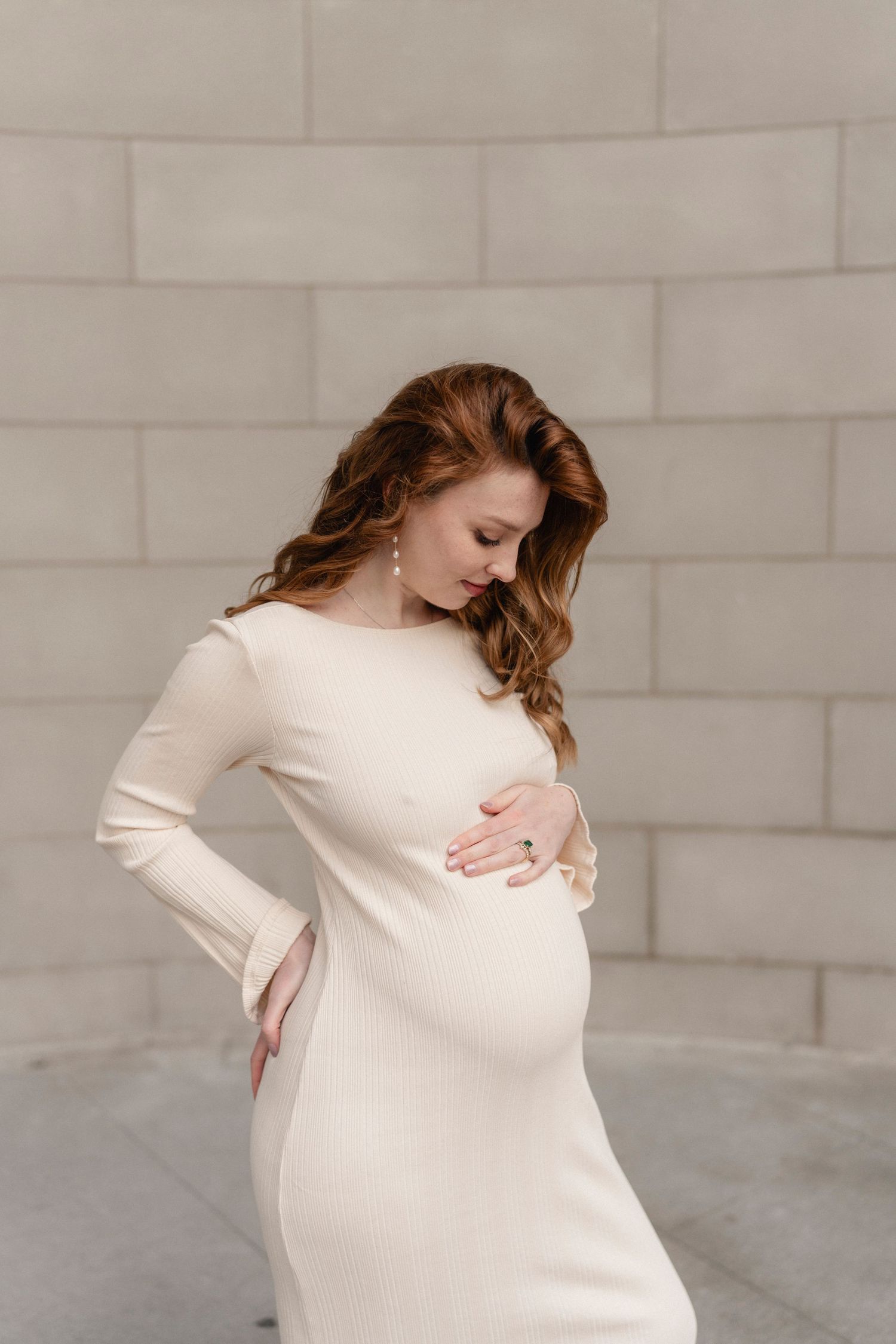 Elegant pregnant woman in cream fitted dress cradling baby bump against stone wall.