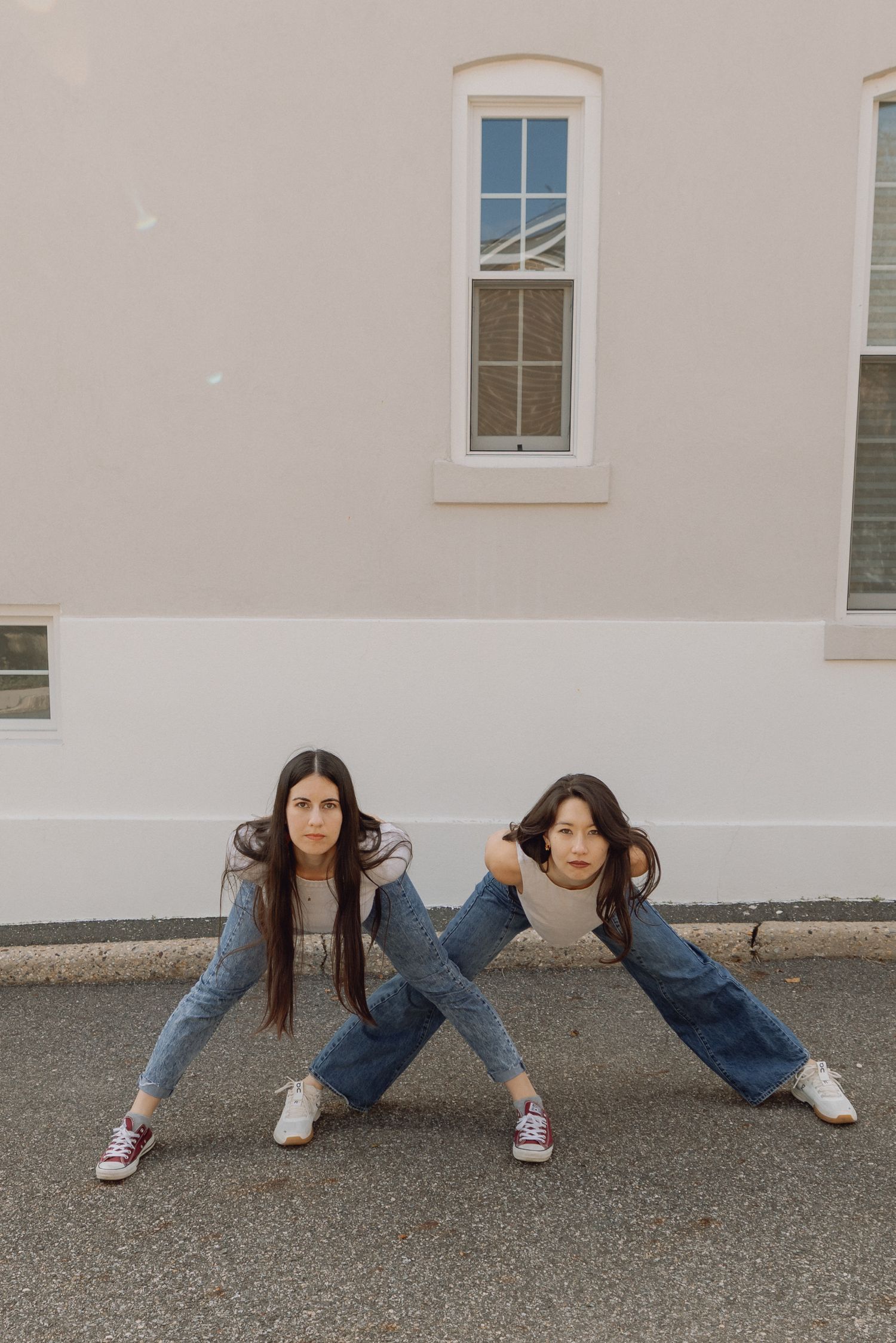 Two young women in jeans pose playfully back-to-back outdoors near a beige building.