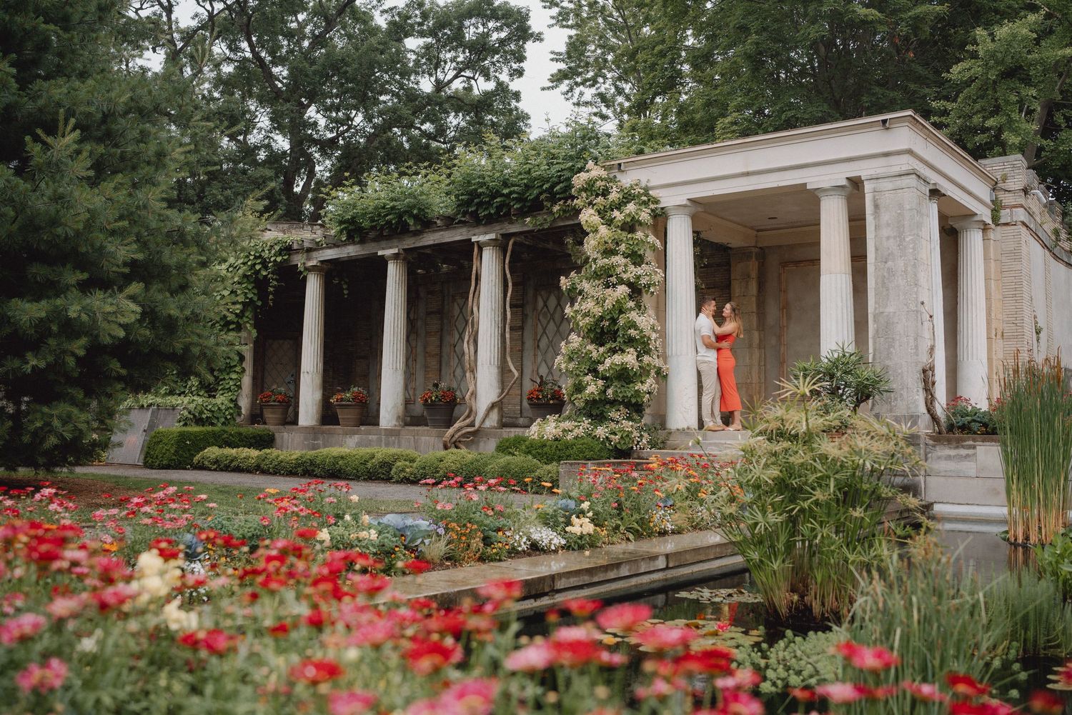 A couple stands on steps of a classical garden pavilion surrounded by vibrant red flowers.