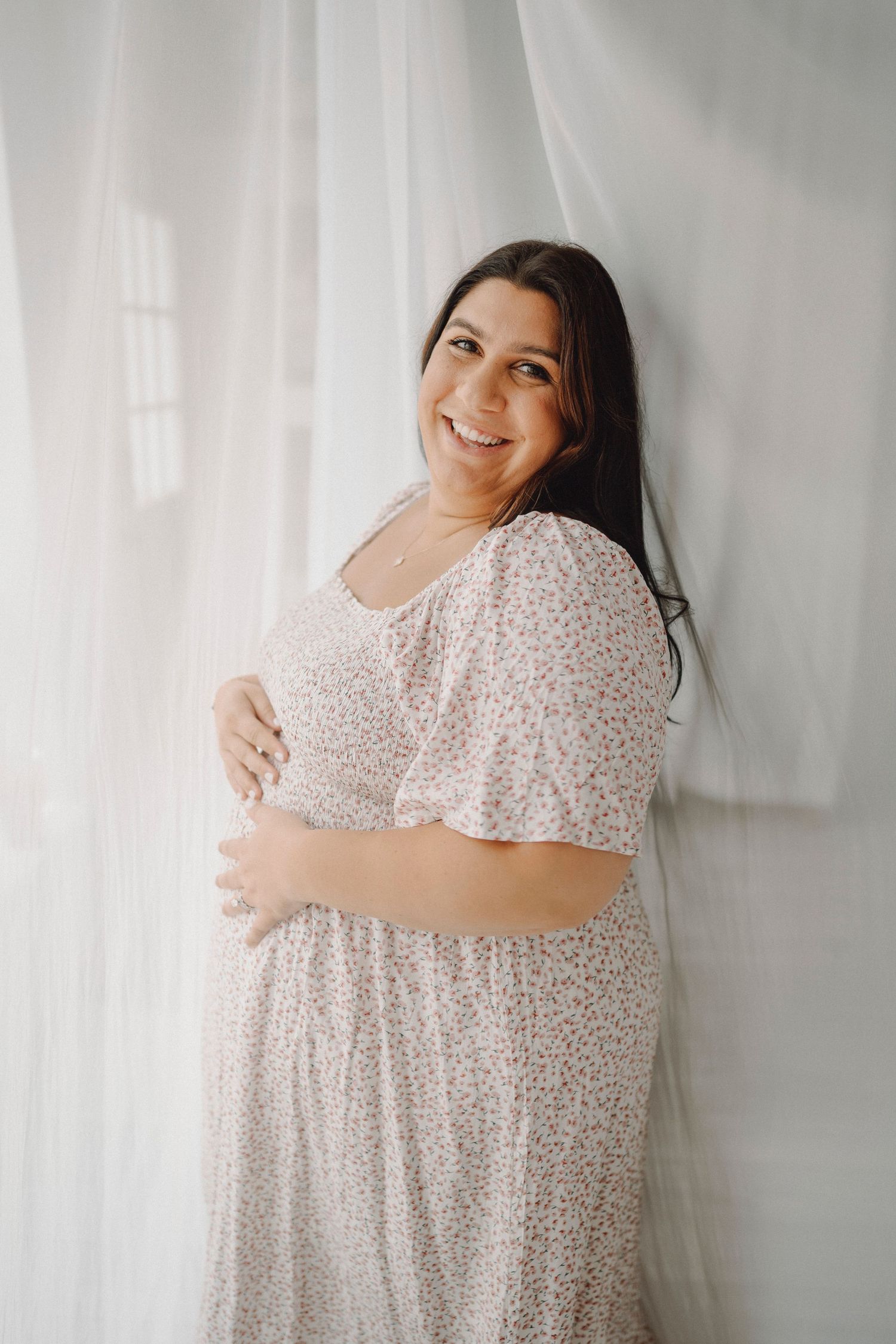 Smiling pregnant woman in floral dress by white curtains.