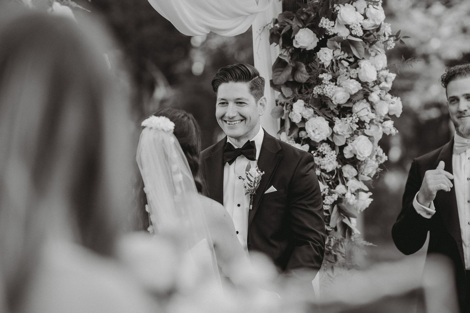 Black and white photo of groom smiling joyfully as bride approaches at outdoor wedding ceremony.