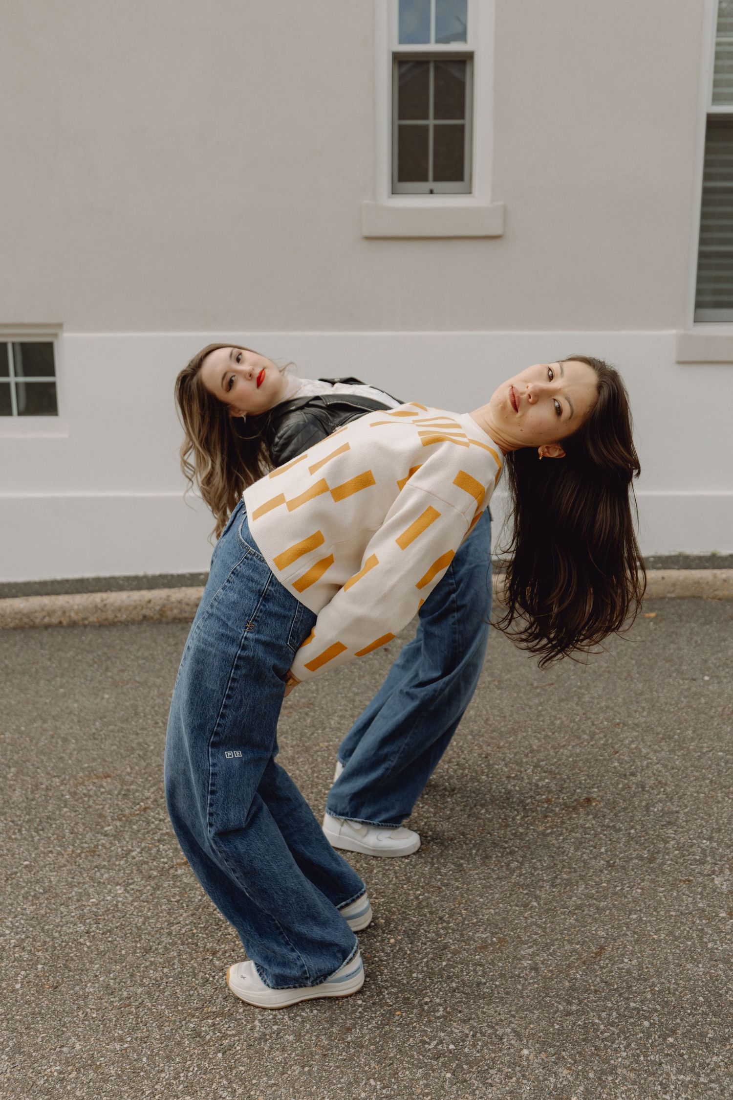 Two young women in wide-leg jeans and casual tops pose back-to-back outdoors near a white building.