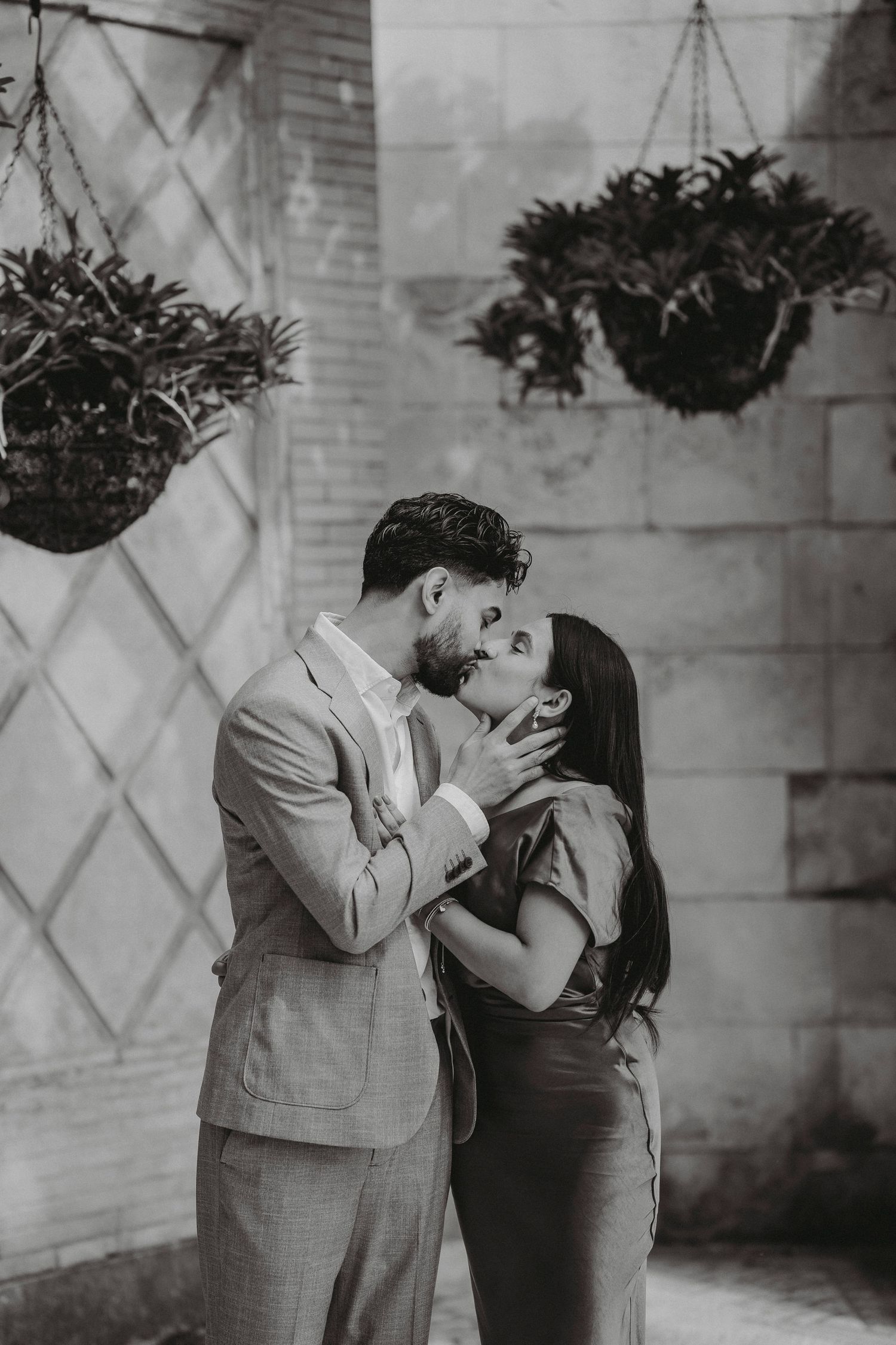 Black and white photo of couple sharing a tender kiss beneath hanging plants on a stone wall.