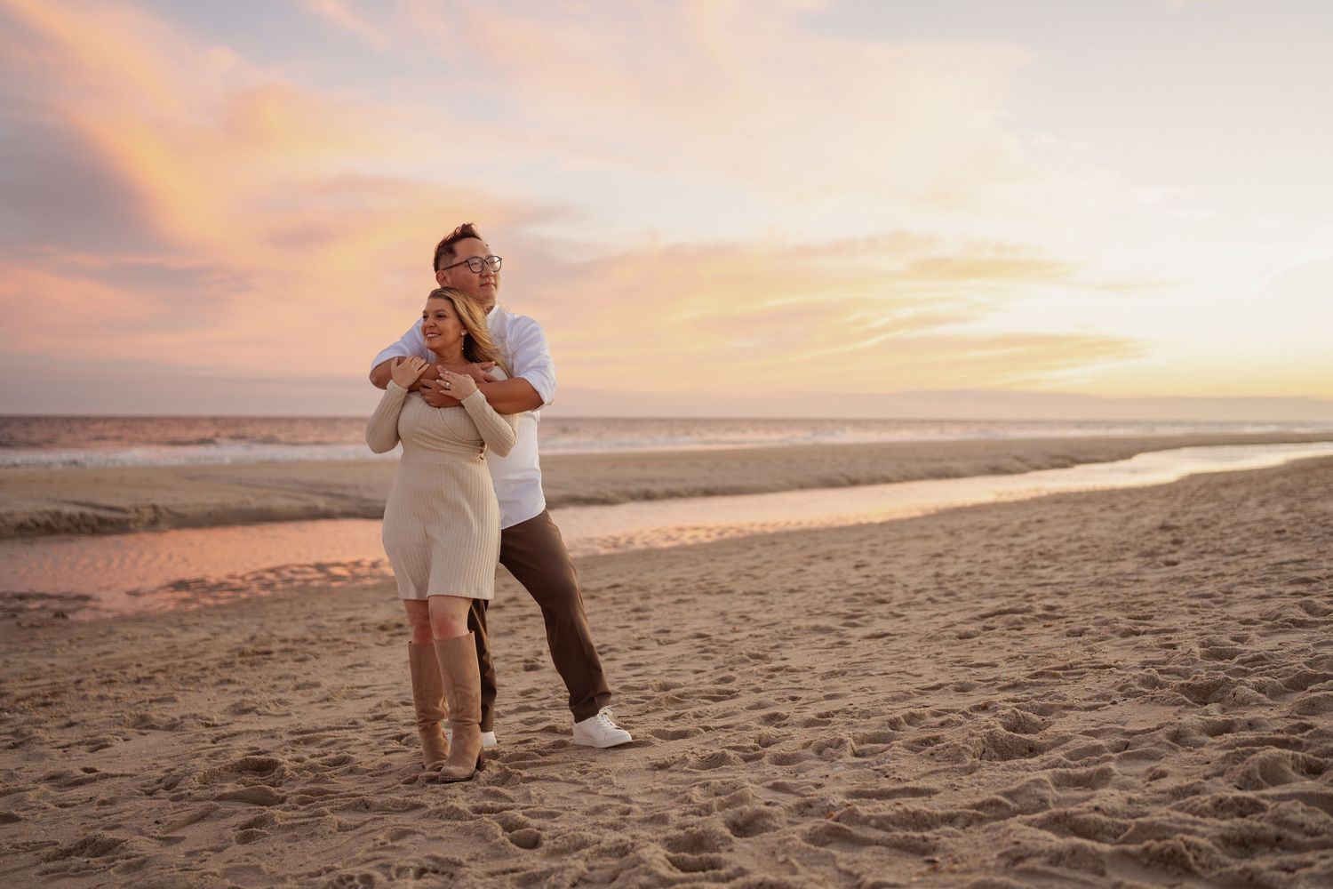 Couple embracing on a sandy beach at sunset with soft pink and orange clouds over the calm ocean waves.