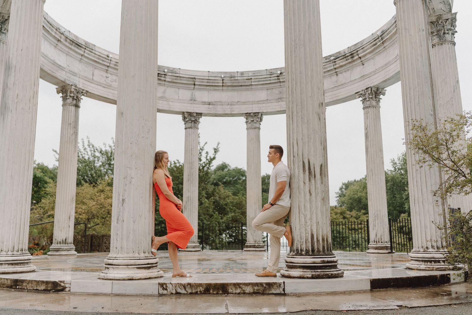 Couple in coral dress and casual outfit lean against marble columns inside a circular neoclassical rotunda surrounded by green trees.