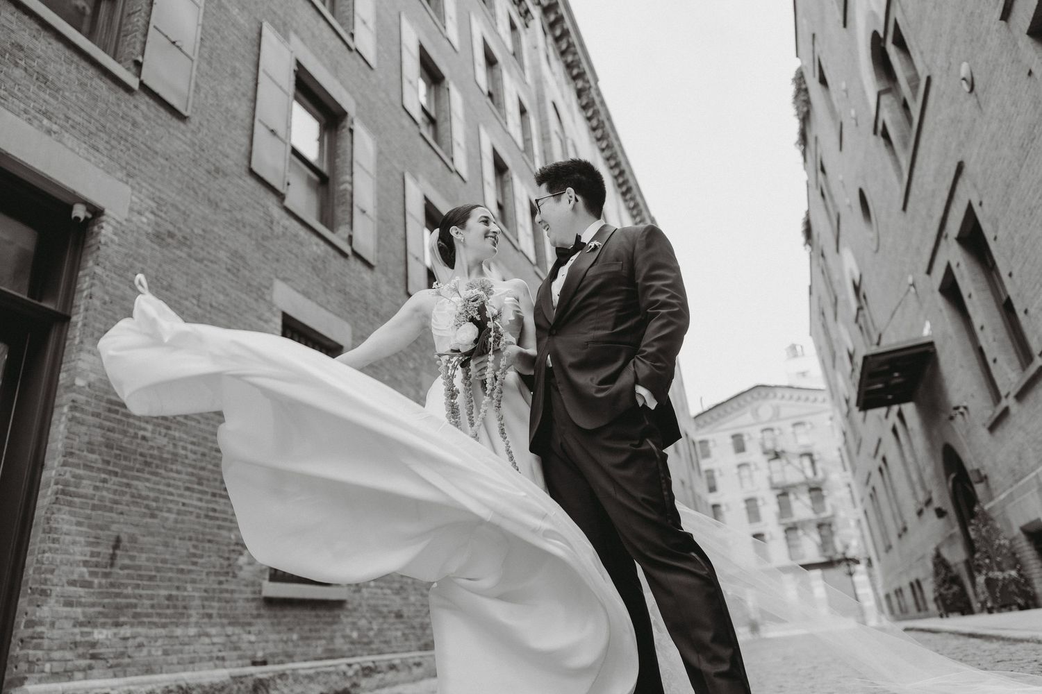 Bride and groom share a romantic moment in a city alley, wedding dress flowing dramatically in the wind.