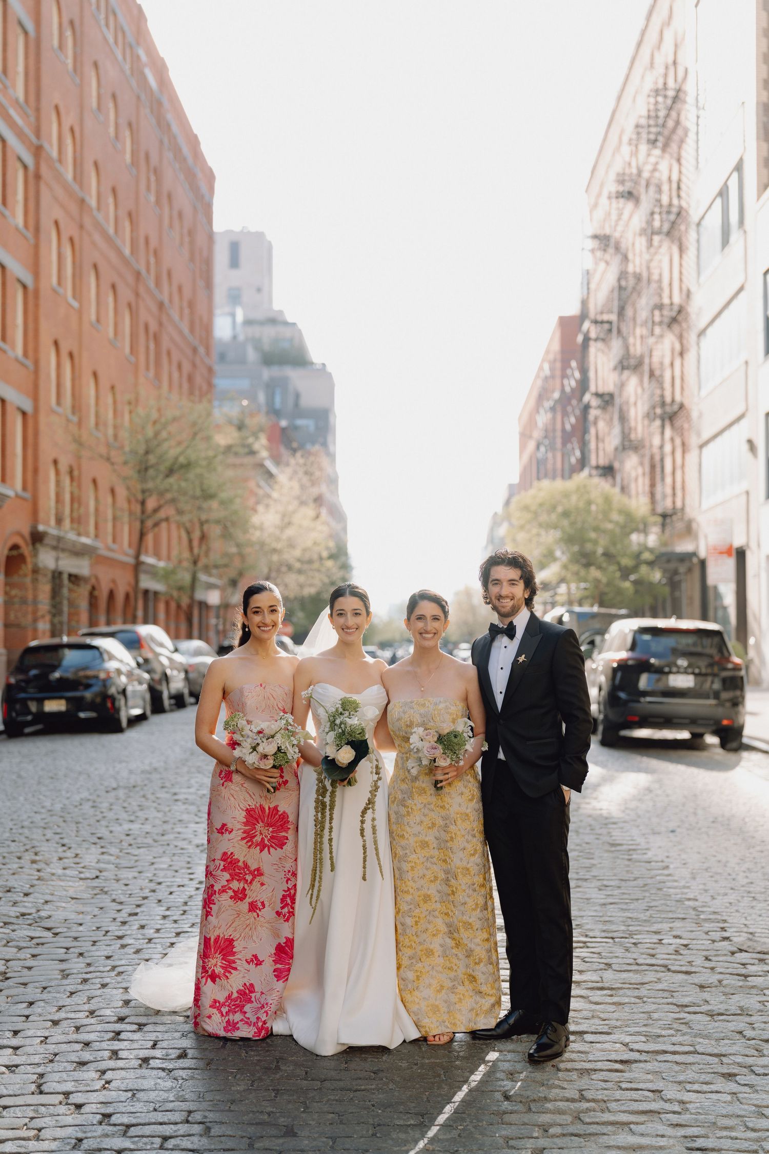 Bride with two bridesmaids in floral gowns and groomsman pose on cobblestone city street.