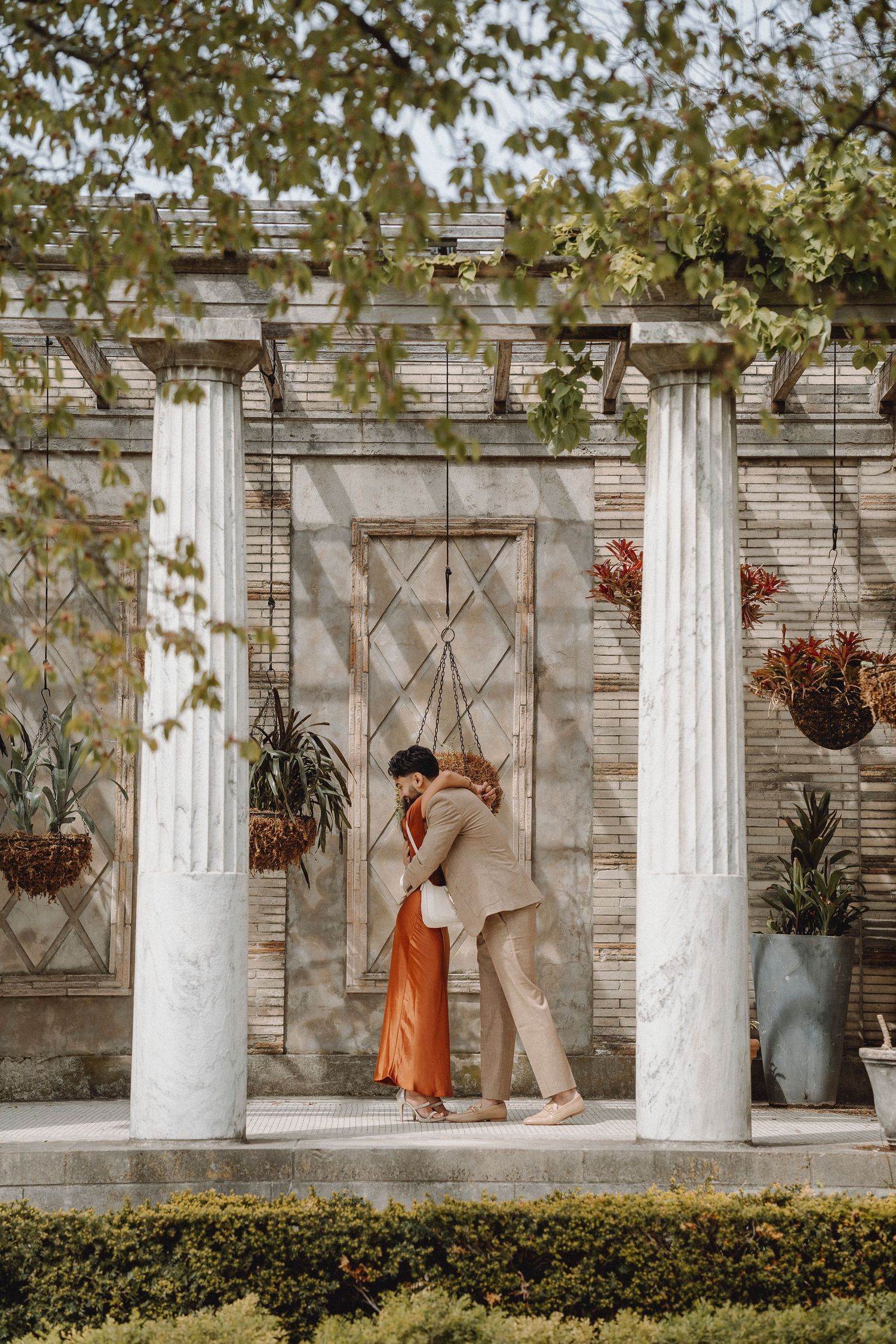 Couple shares a romantic embrace between white marble columns of an ornate garden structure.