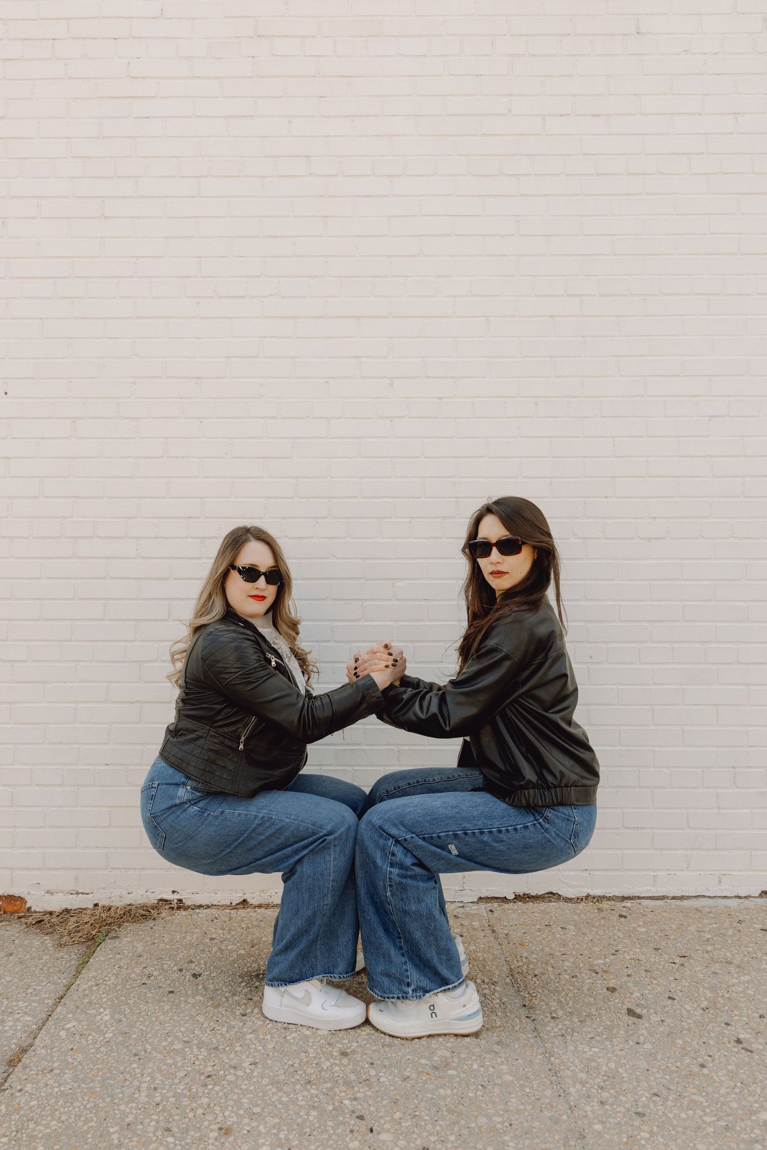 Two women in matching black leather jackets and jeans squatting while holding hands against white brick wall.