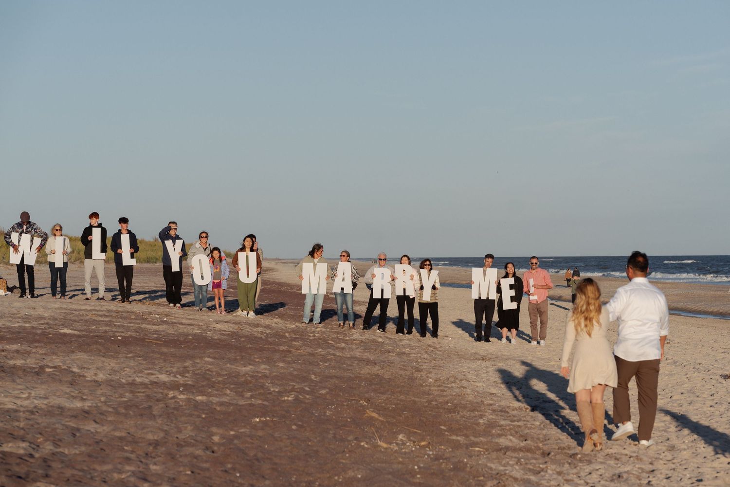 Wedding guests in light-colored attire stand in two lines on a sandy beach as a bride and groom walk toward the ocean at sunset.