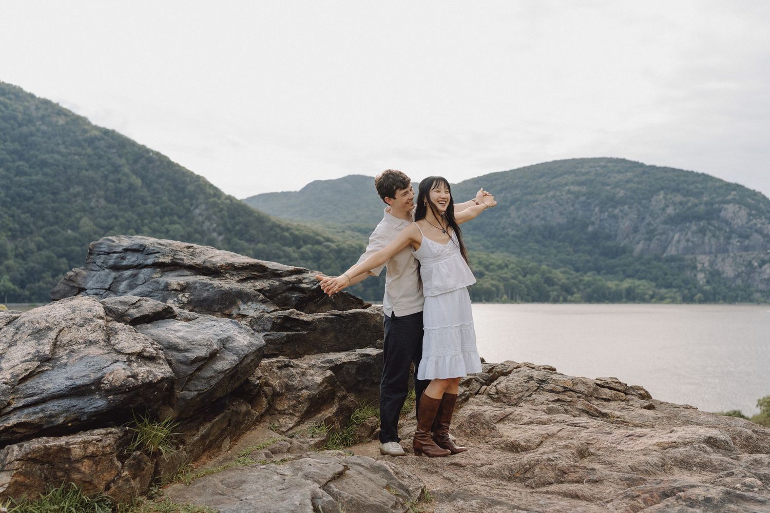 Couple standing with arms outstretched on a rocky overlook above a wide river, with green forested mountains in the background.