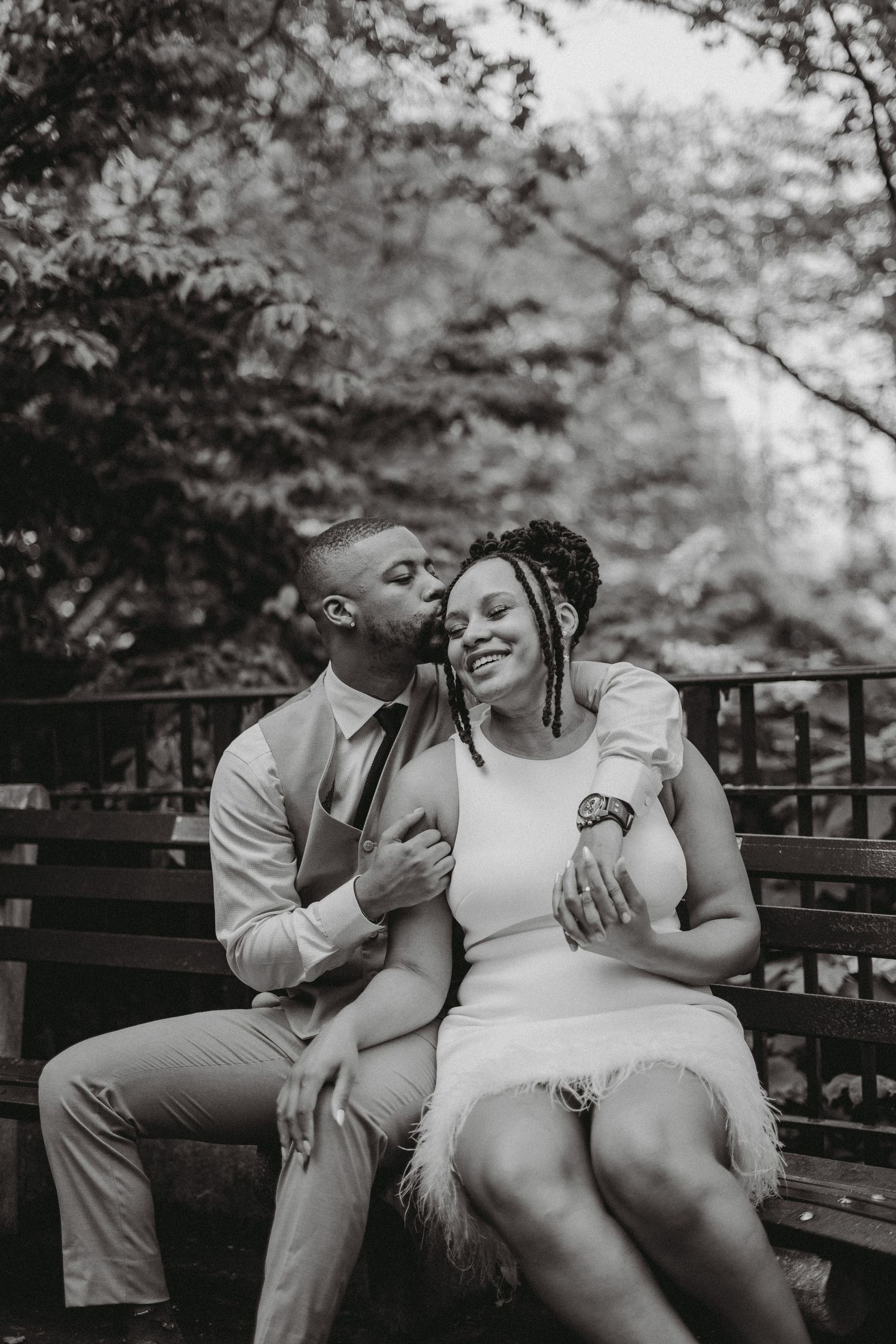 Black and white photo of a couple sitting on a bench, man kissing woman's cheek as she smiles joyfully.