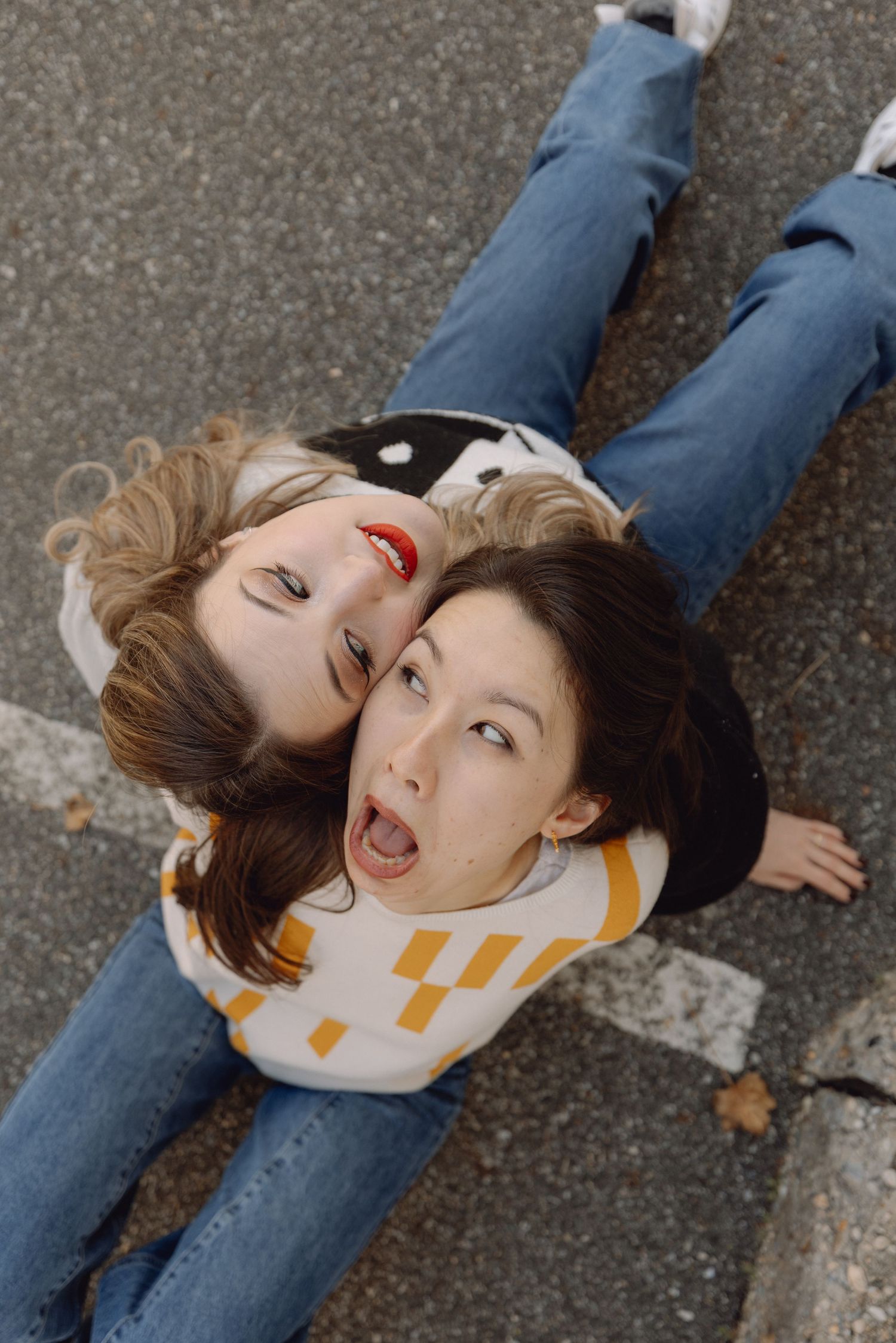 Two young women lying on pavement looking up at camera with playful expressions and wide-leg jeans.