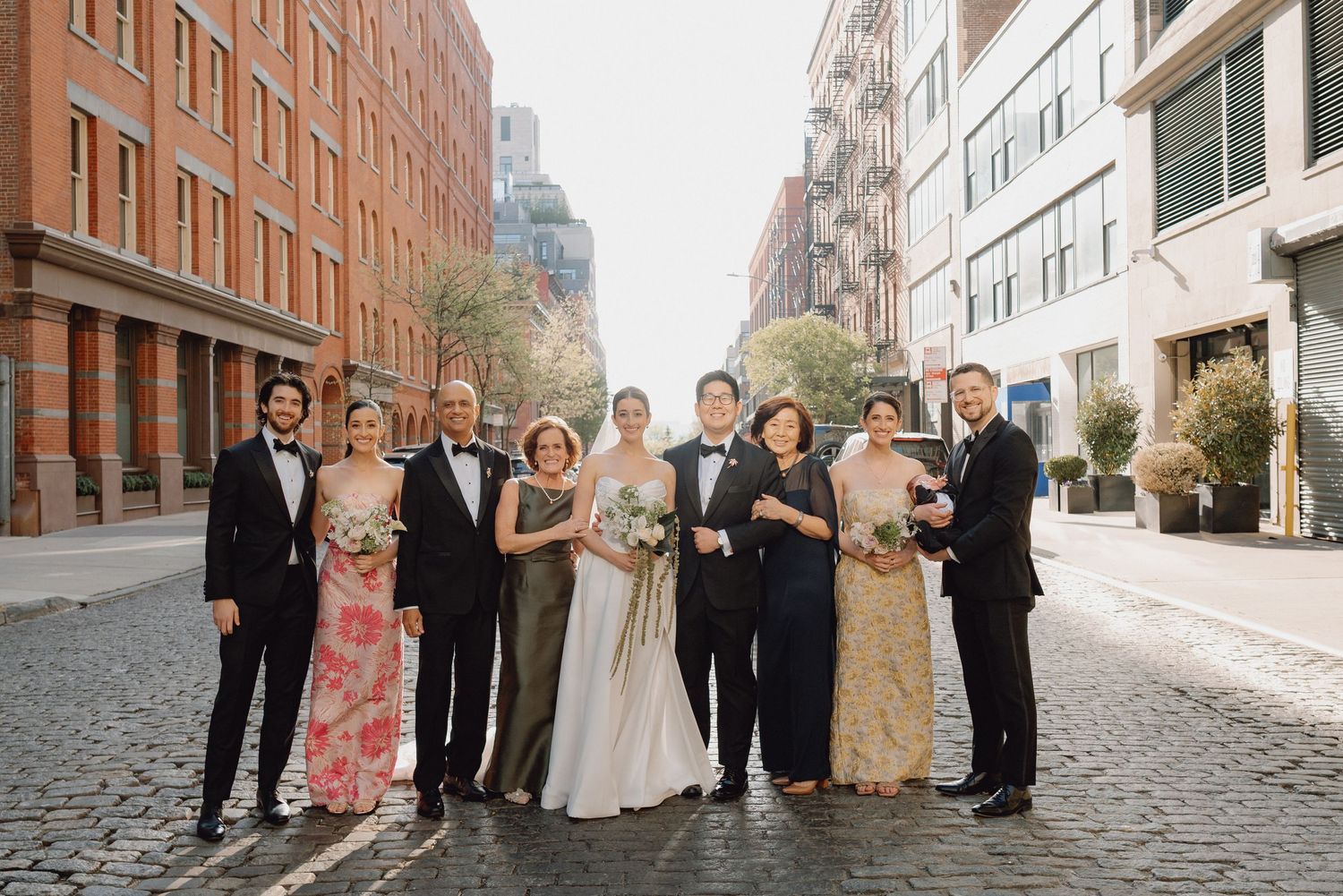 Wedding party and families pose together on sunlit cobblestone street in New York City.