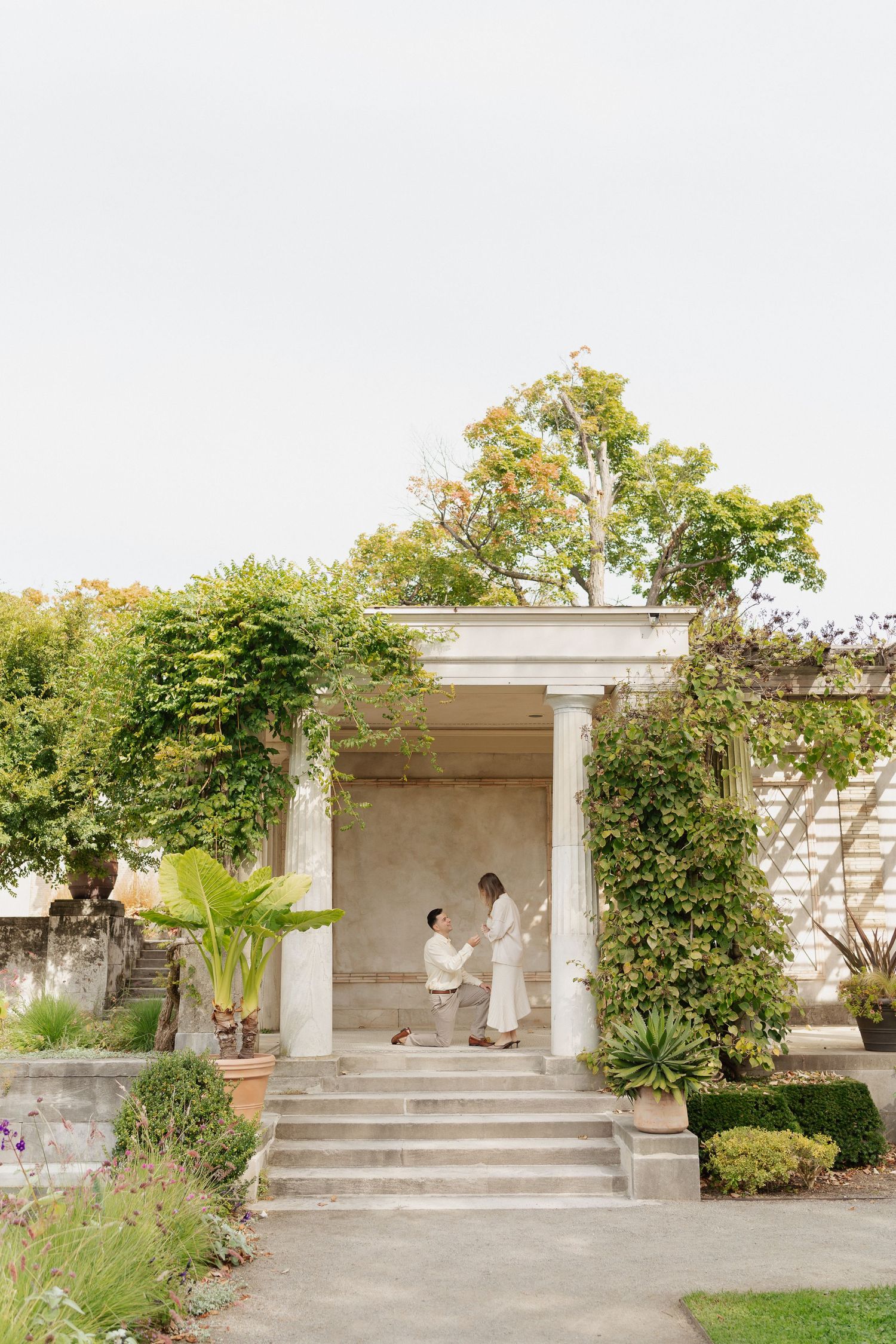 Man kneeling on one knee proposing to a woman beneath a white columned garden pavilion with climbing vines and lush greenery.