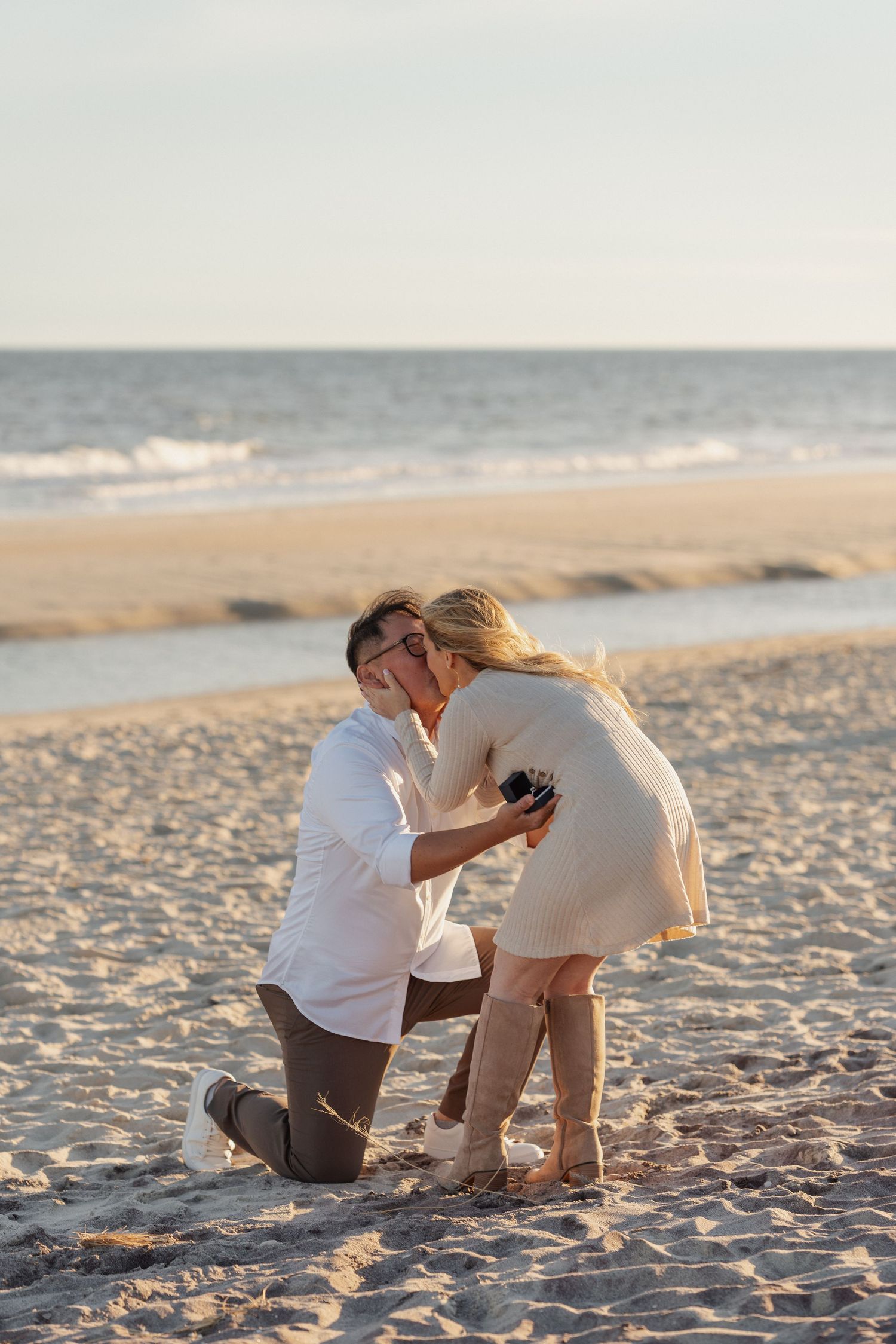 Man kneeling on one knee holding a ring box as a woman in a cream dress and tall boots covers her face during a beach proposal at sunset.
