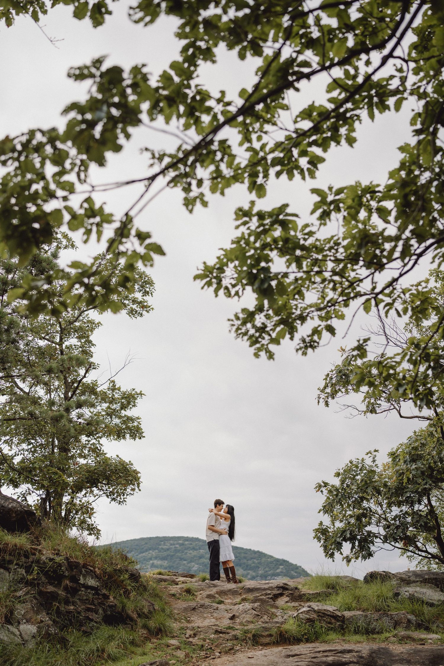 Couple embracing on a rocky hilltop framed by green tree branches, with a distant mountain and overcast sky behind them.