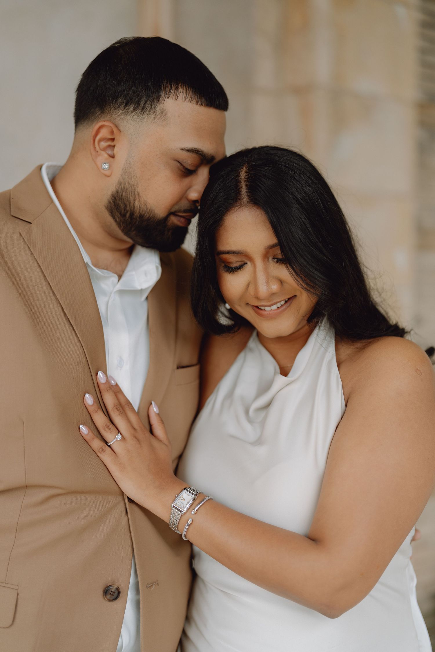 Engaged couple in elegant attire sharing an intimate moment, man in tan suit, woman in white dress.
