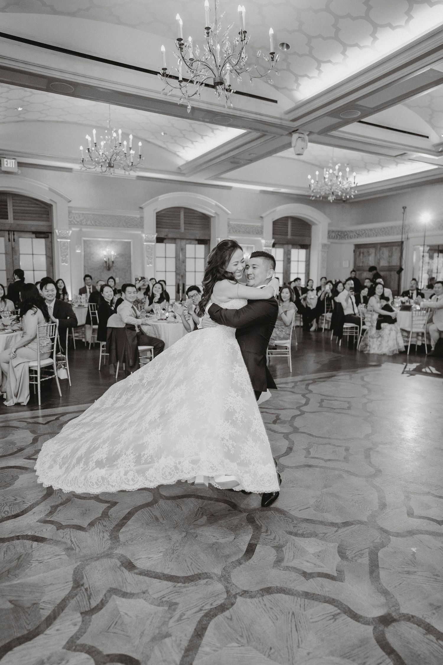 Bride and groom share their first dance in a grand ballroom in black and white photo.