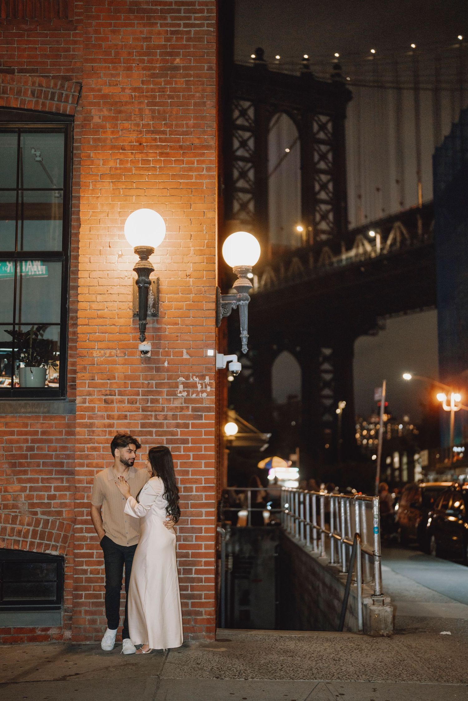 Couple leaning against brick wall under warm lights with Manhattan Bridge at night.