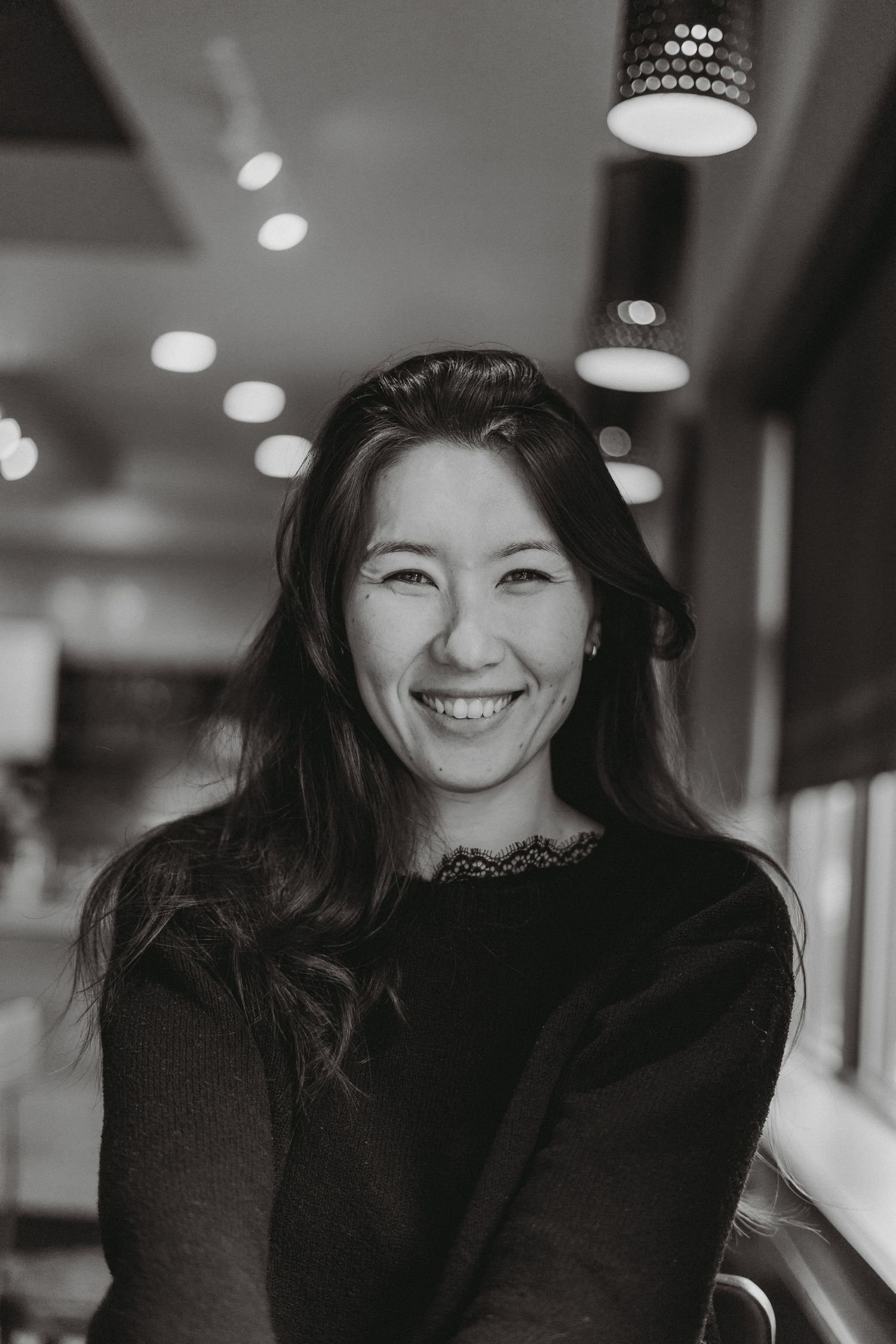 Black and white portrait of a smiling woman with long dark hair in a softly lit cafe setting.