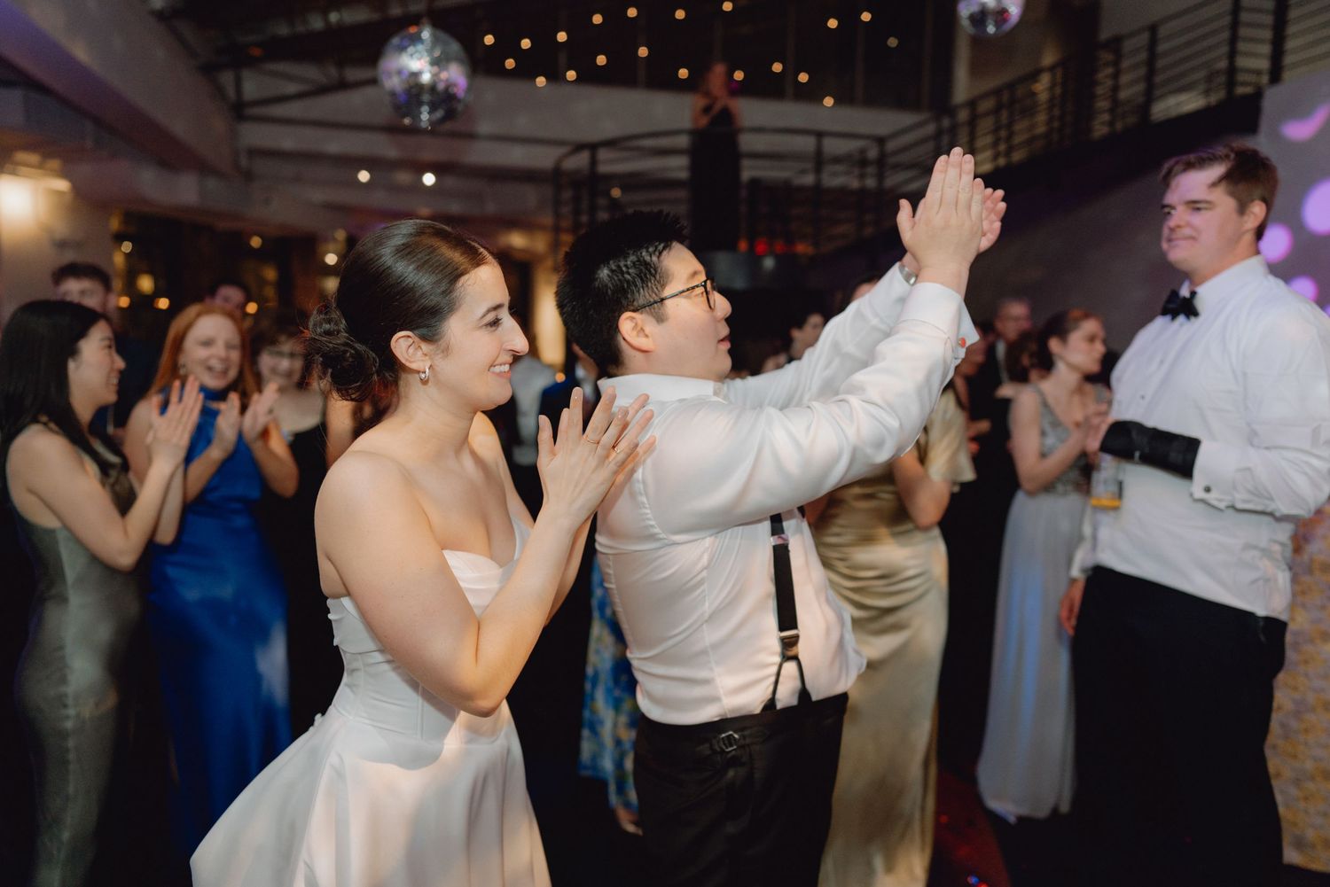Bride and groom dance joyfully together as wedding guests clap and celebrate around them.