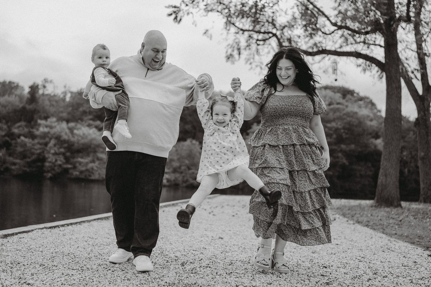Black and white photo of a joyful family walking and swinging their daughter between them outdoors.