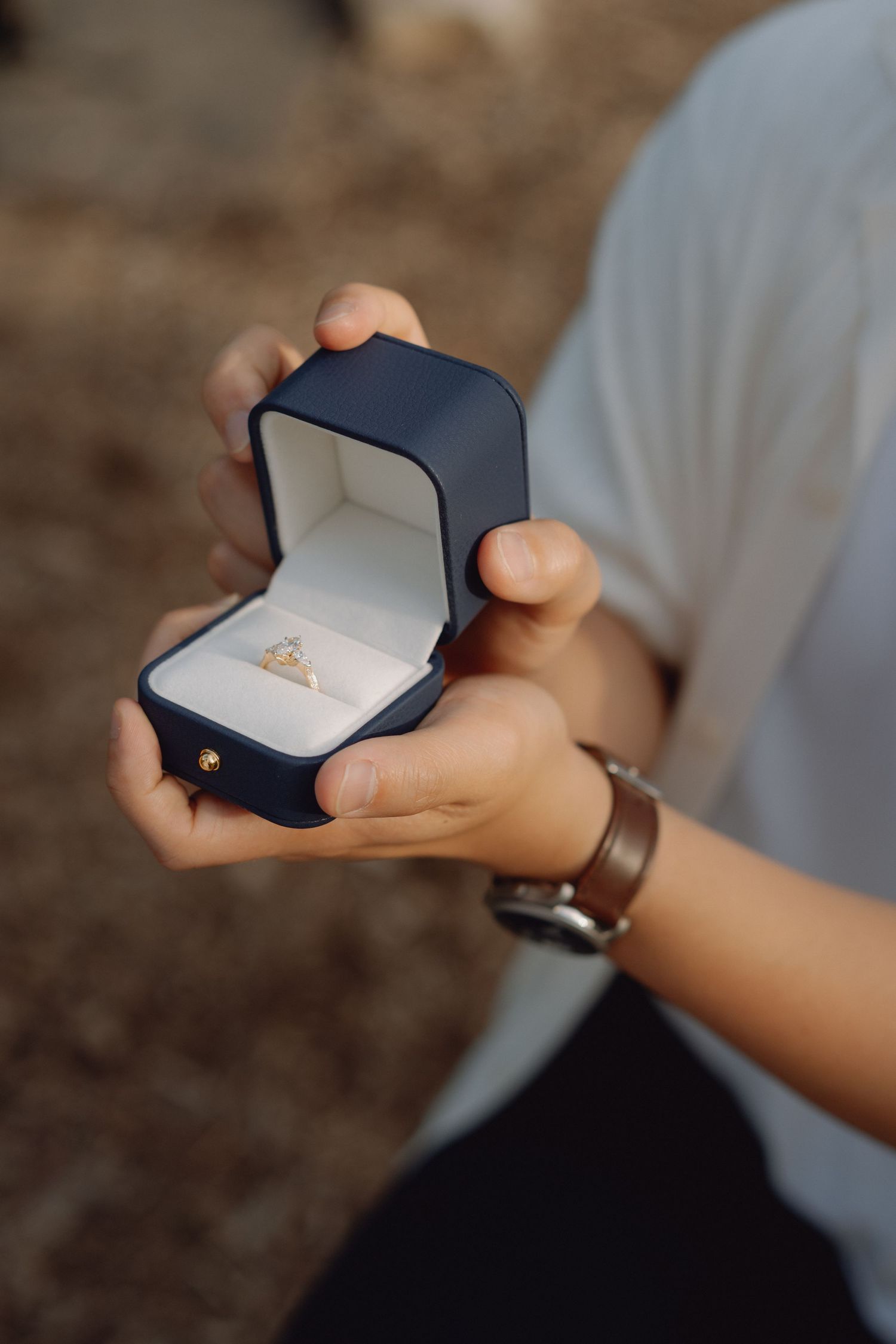 Hands holding an open navy blue ring box with a diamond engagement ring inside, worn with a leather strap watch and white shirt.