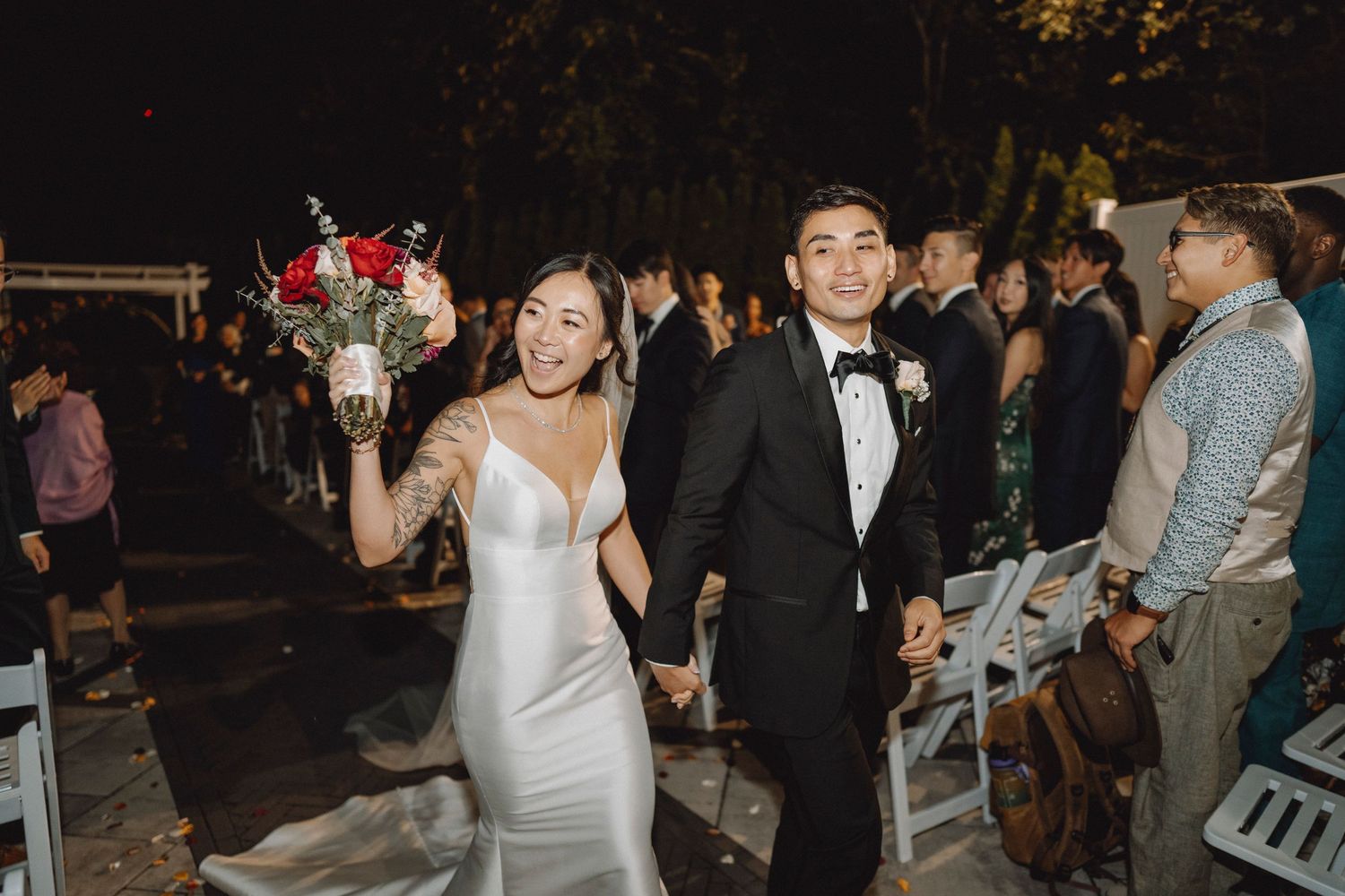 Bride and groom walk down the aisle smiling, bride holding red and white floral bouquet at night.