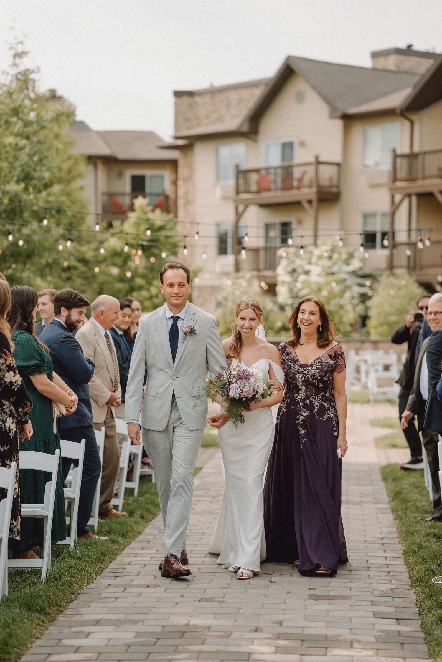 Bride in white gown walks down brick aisle with escorts during outdoor wedding ceremony.