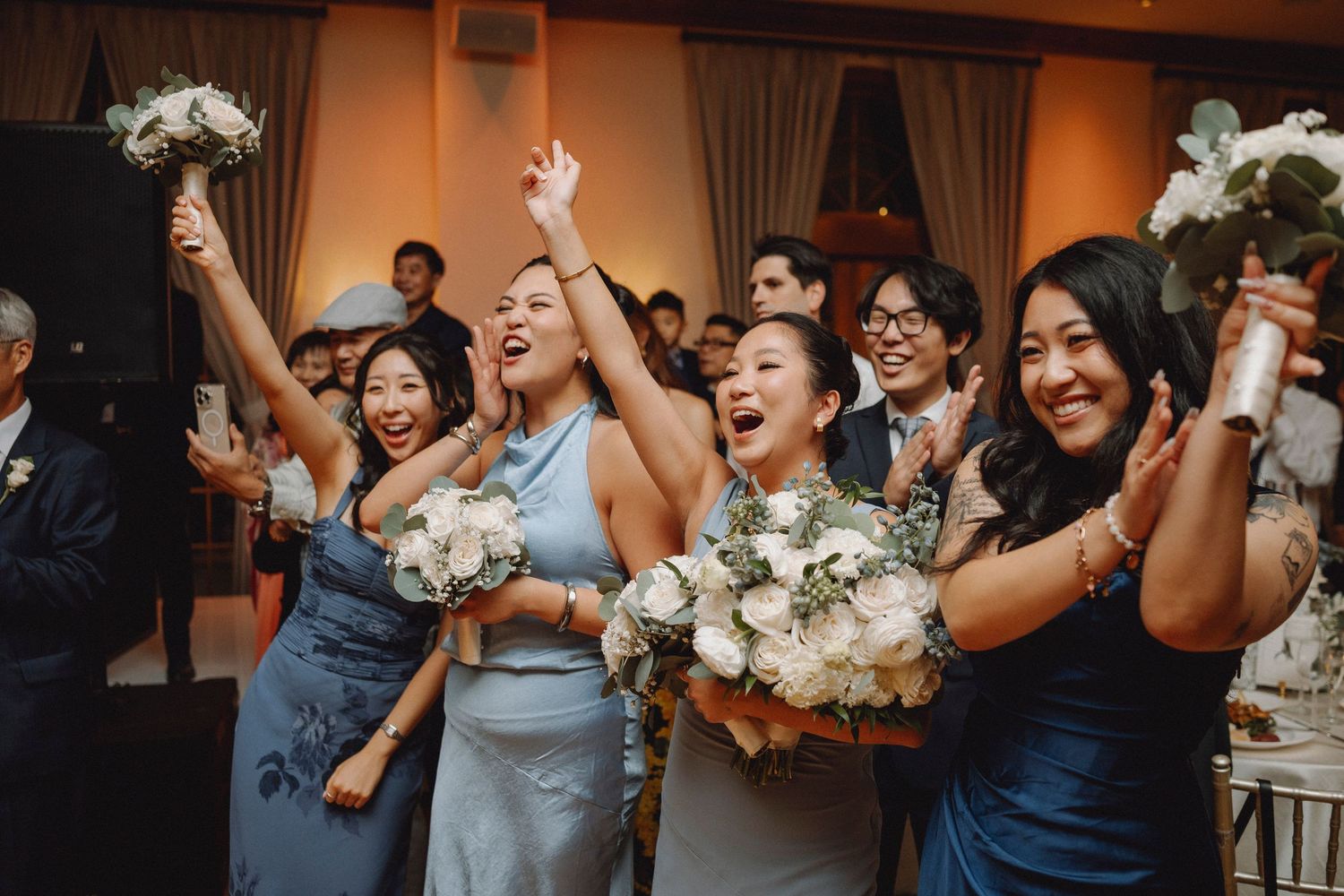 Excited bridesmaids in blue dresses celebrate with bouquets raised at elegant wedding reception.