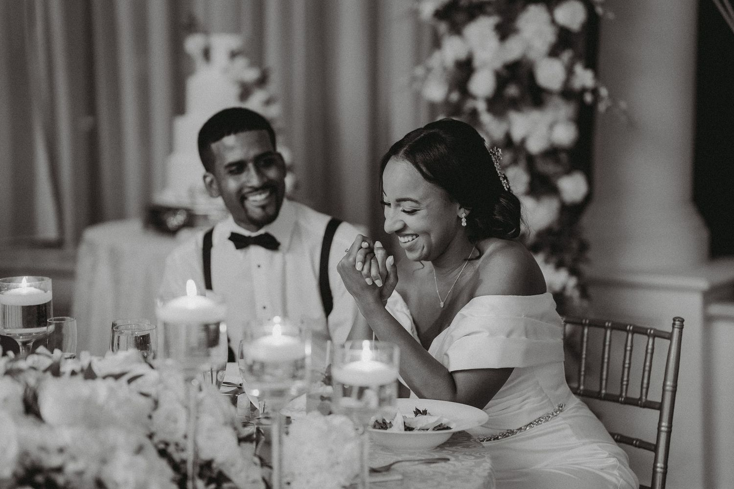 Bride and groom laugh joyfully at sweetheart table surrounded by candles and floral arrangements.