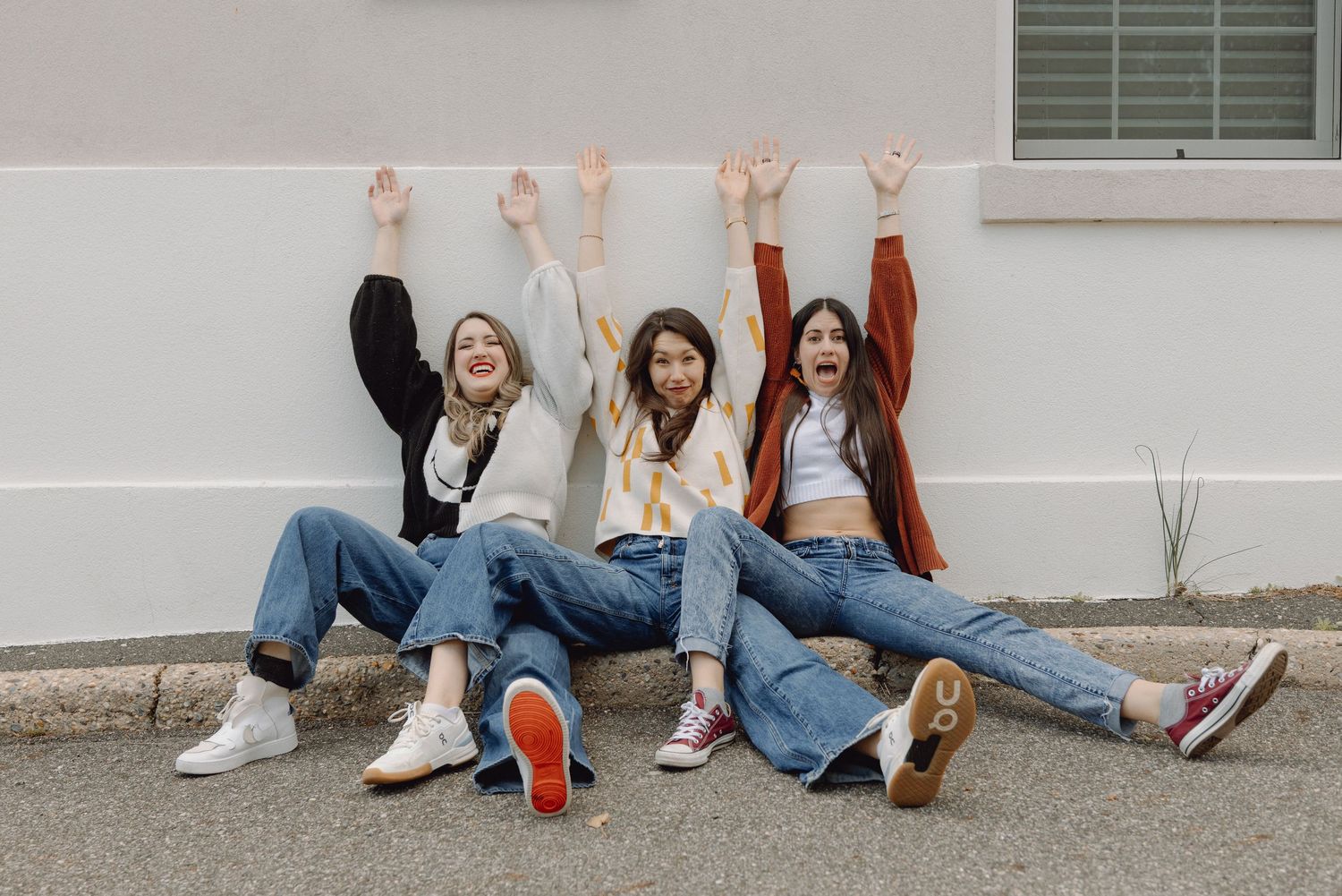 Three young women sitting against a wall with arms raised, wearing jeans and sneakers, laughing and celebrating outdoors.