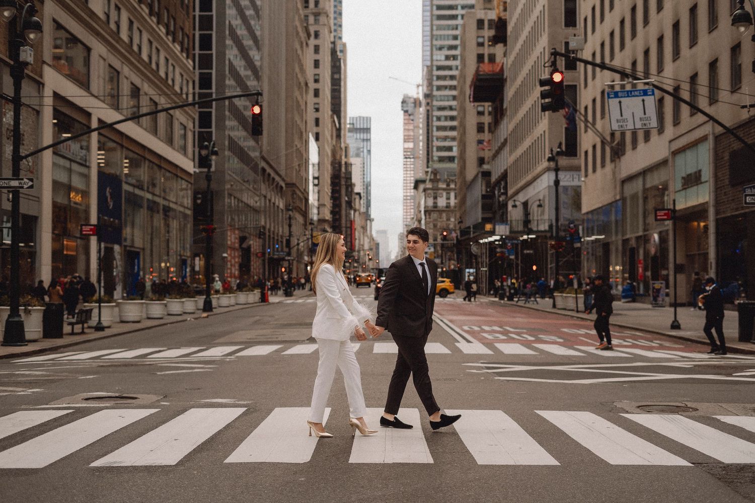 Couple holding hands crossing a zebra crossing in a busy city street surrounded by tall buildings.