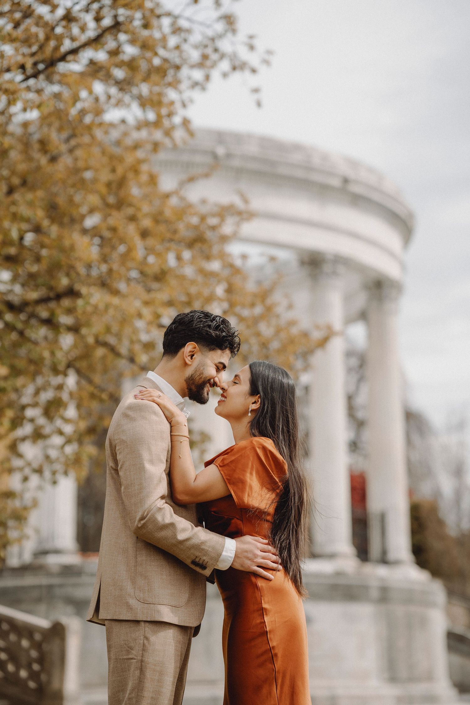 Romantic couple nearly kissing in front of a classical white columned rotunda during fall season.