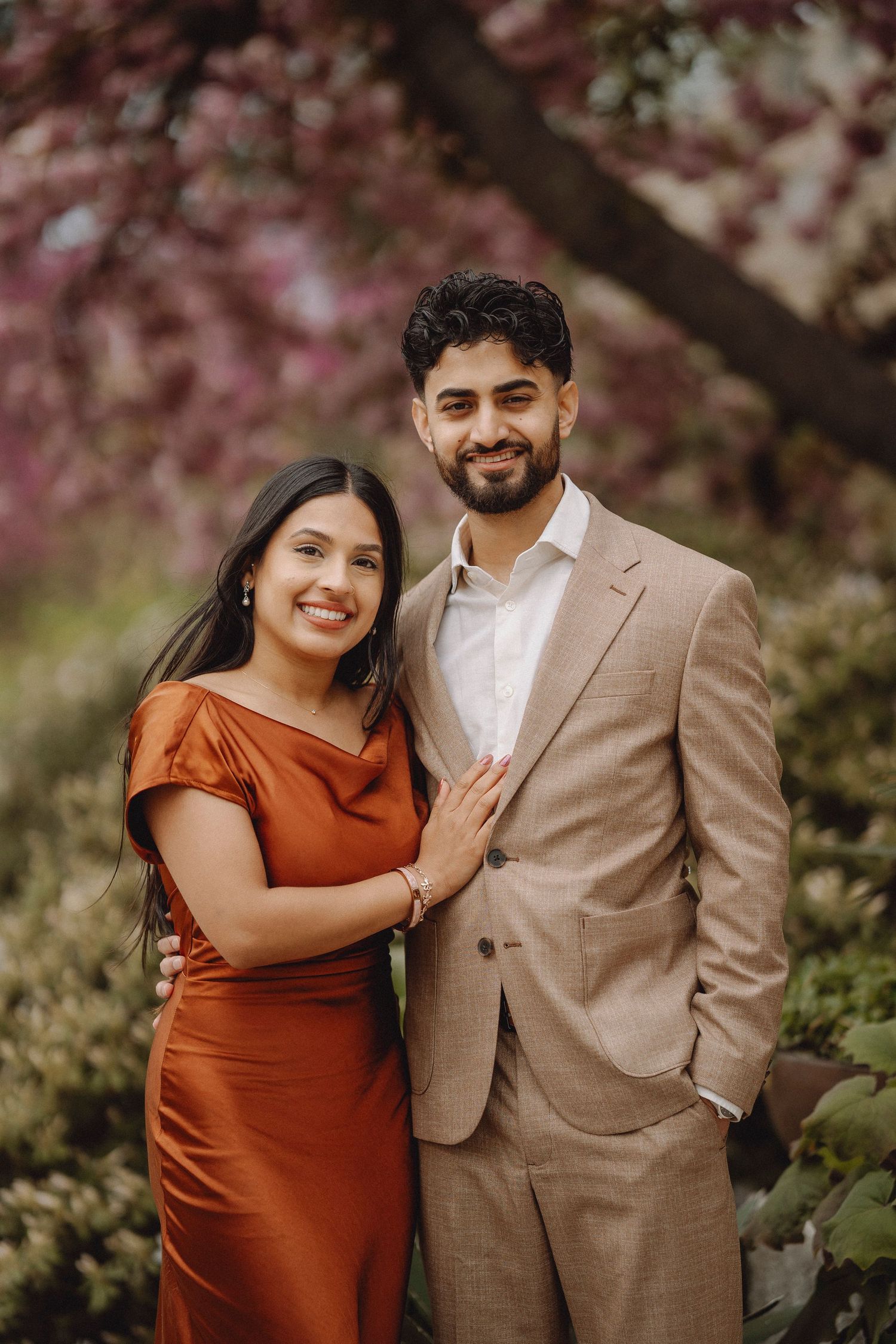 Smiling couple posing under blooming pink cherry blossom tree, woman in rust dress, man in beige suit.