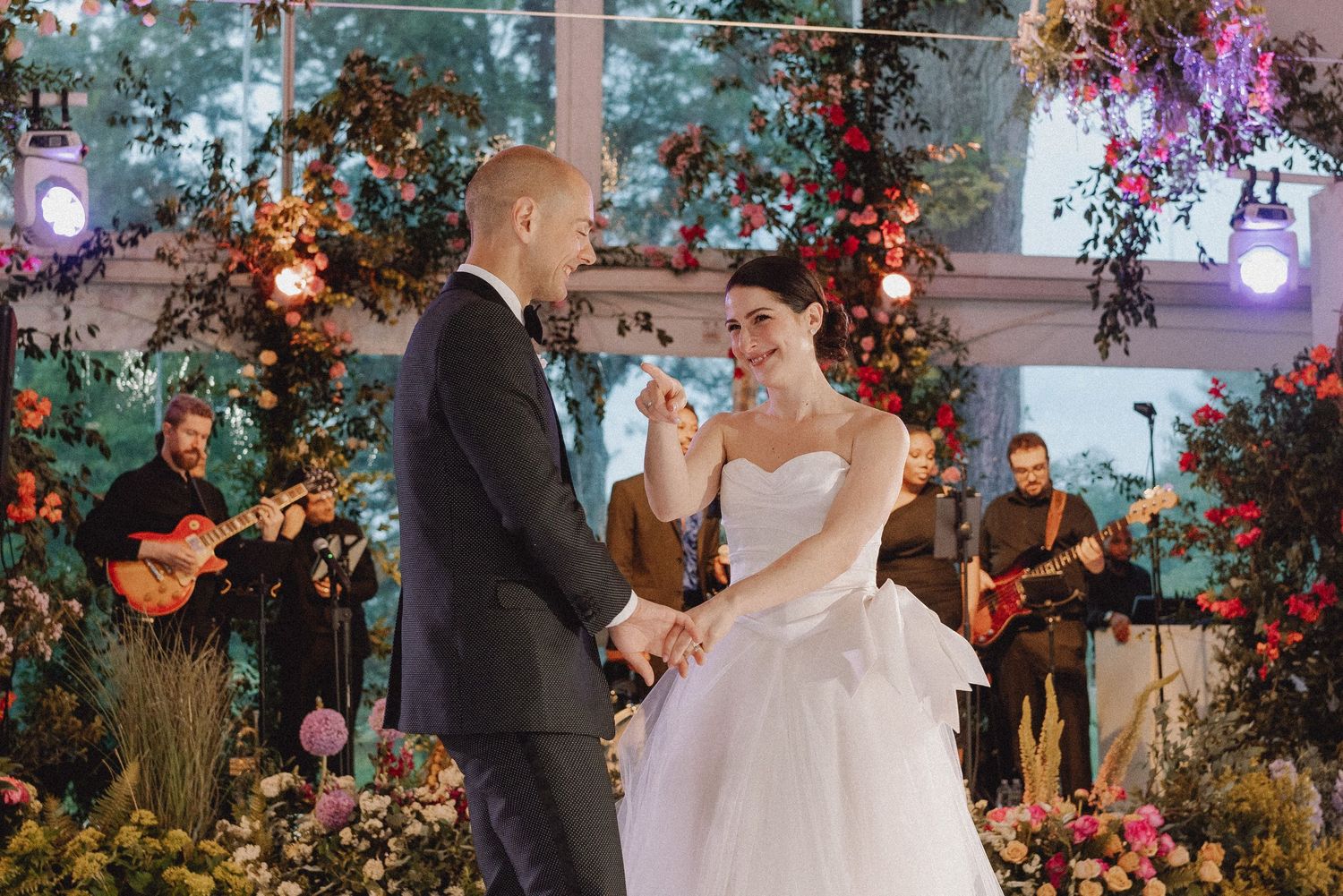 Bride and groom share first dance surrounded by lush floral arrangements and live band at elegant wedding reception.