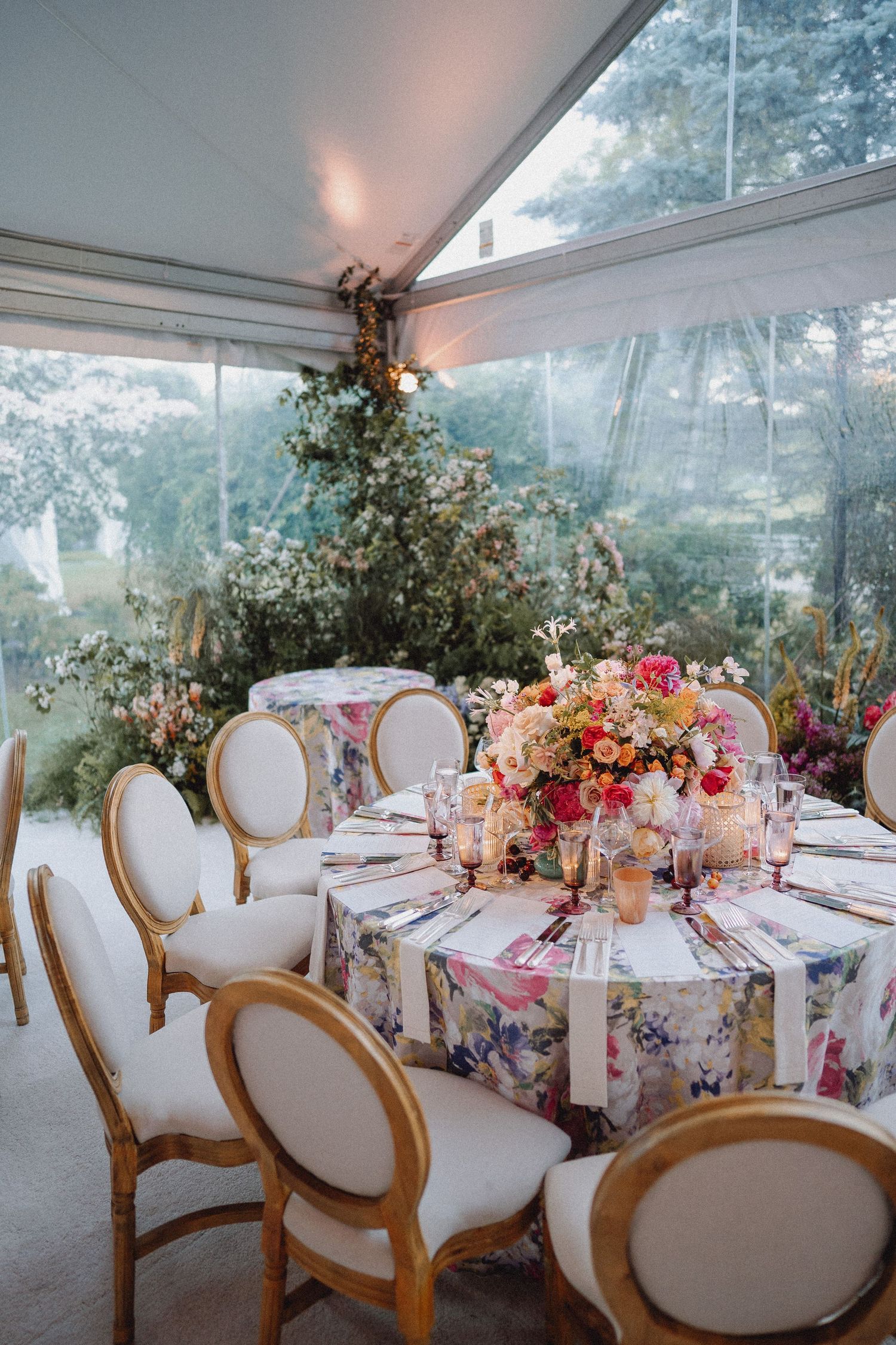 Elegant wedding reception table with floral tablecloth, pink blooms, and gold chairs under a clear tent.