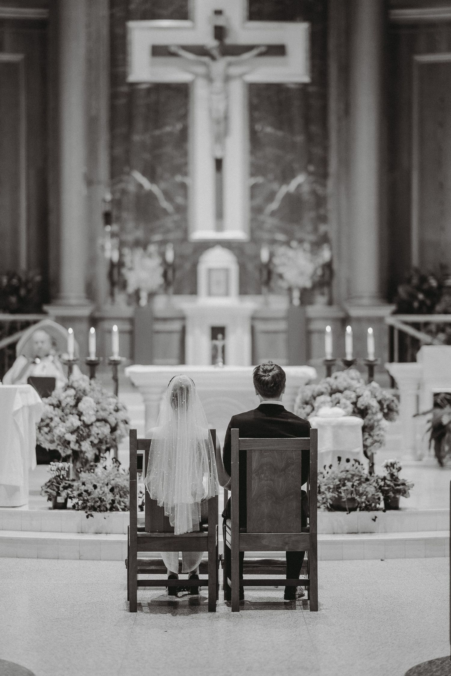 Black and white photo of bride and groom seated before altar with large crucifix and floral arrangements in church ceremony.