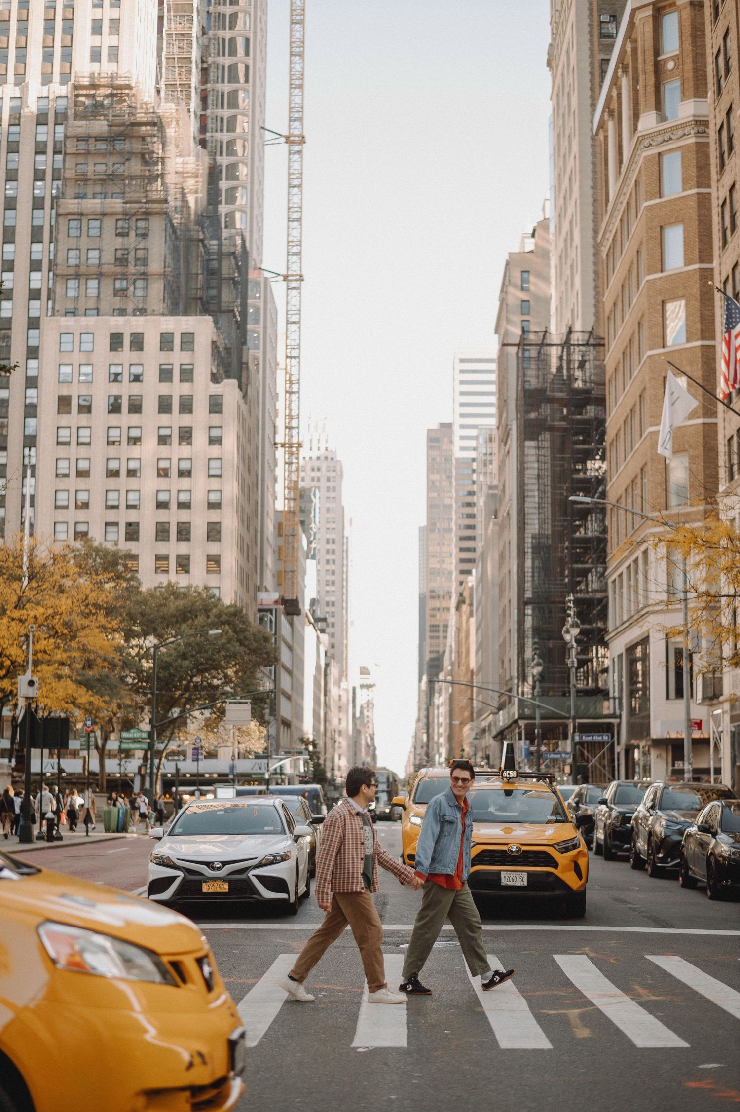 Two men cross a busy New York City street holding hands among yellow taxis and tall buildings.