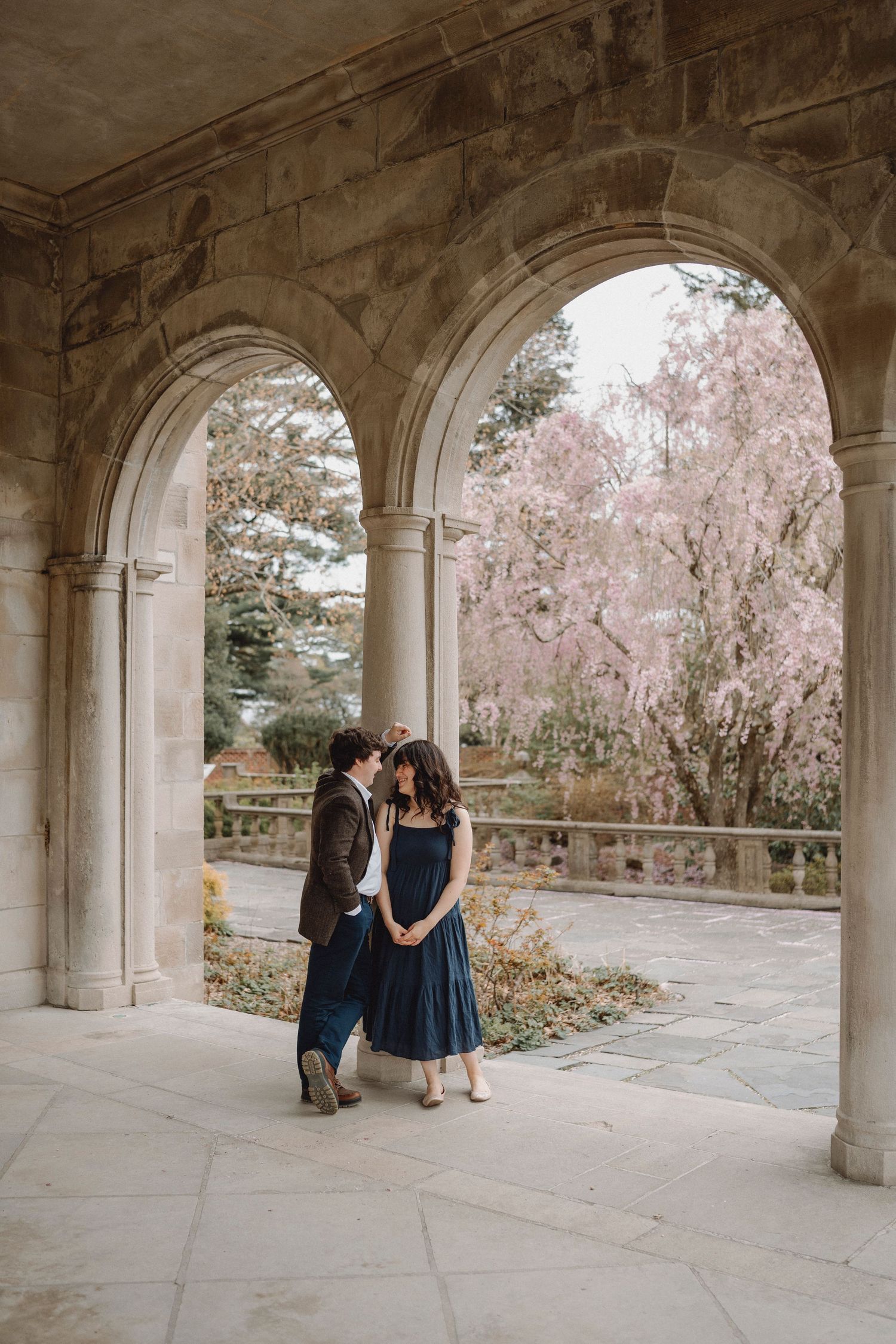 An engaged couple shares a quiet moment under stone arches with pink cherry blossoms behind.