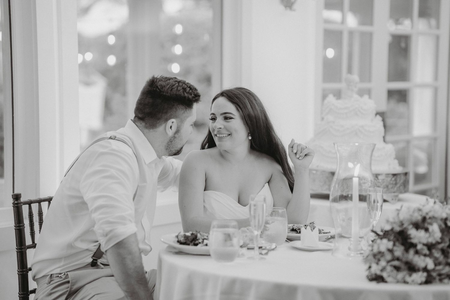 Black and white photo of a bride and groom laughing together at their sweetheart table during reception.